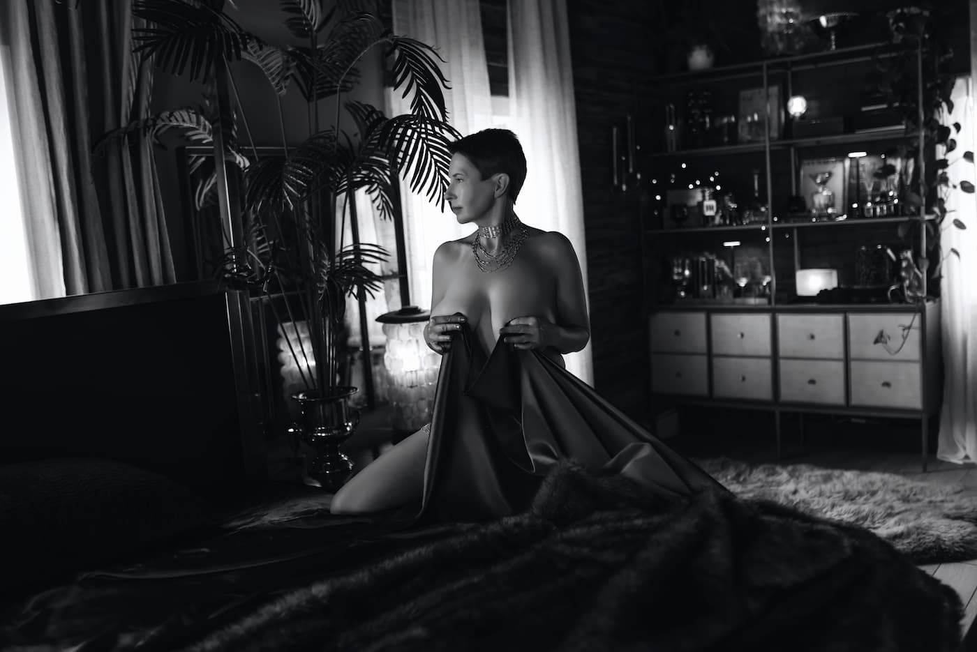 A person sits in moody low-key lighting on a bed, holding a glowing device while surrounded by houseplants and decor.