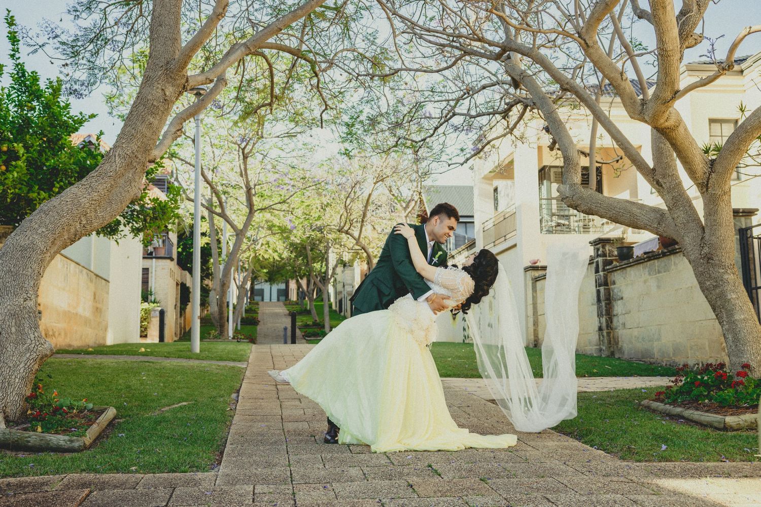 A wedding couple walks along a scenic path lined with trees and buildings on a sunny day.