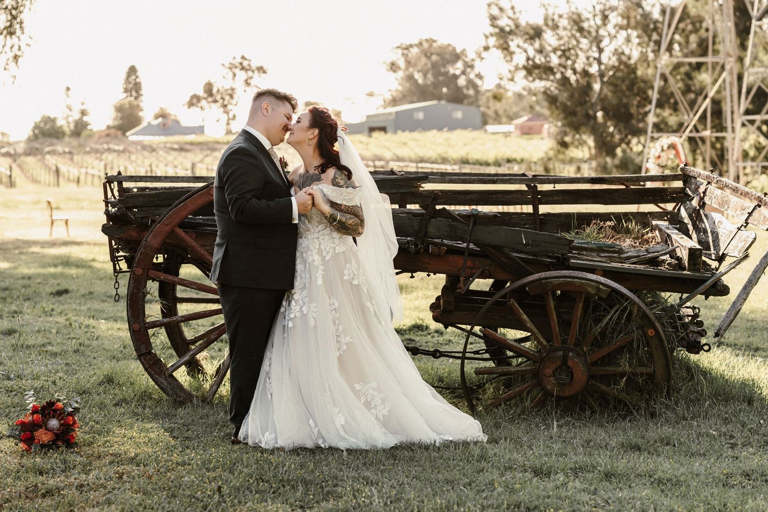 Romantic couple in wedding attire embraces by vintage wooden wagon wheel in rustic outdoor setting at sunset.