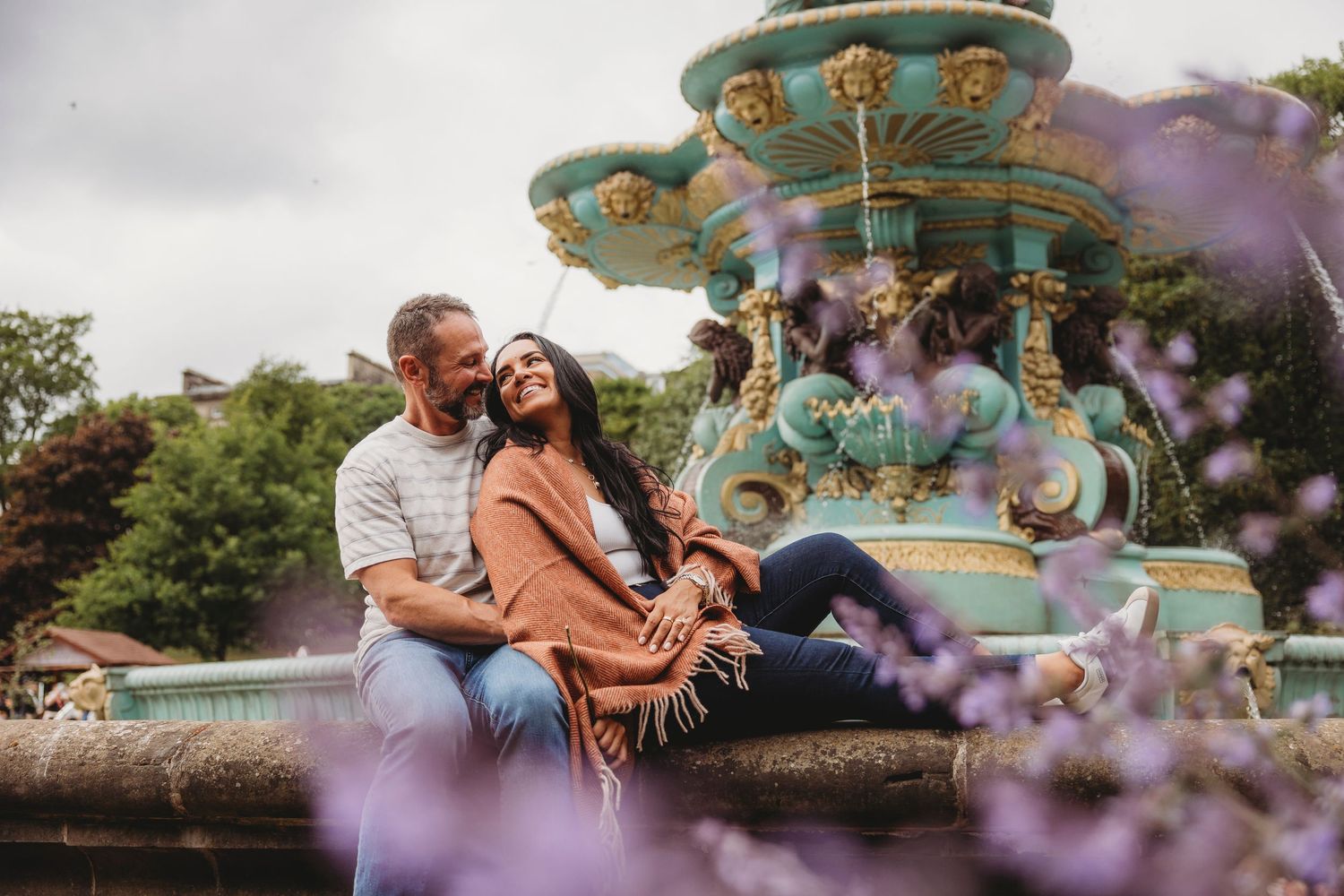 A romantic couple sits together near a vintage carousel with purple flowers in the foreground at an amusement park.