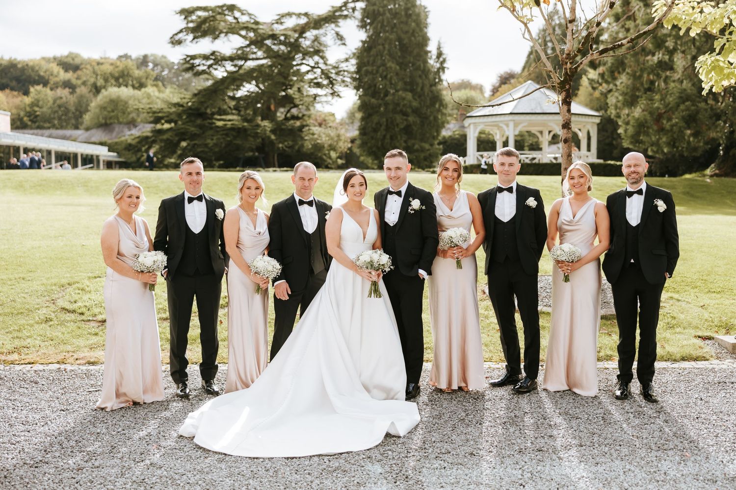 Wedding party in formal attire poses together on a sunny lawn at an outdoor celebration.