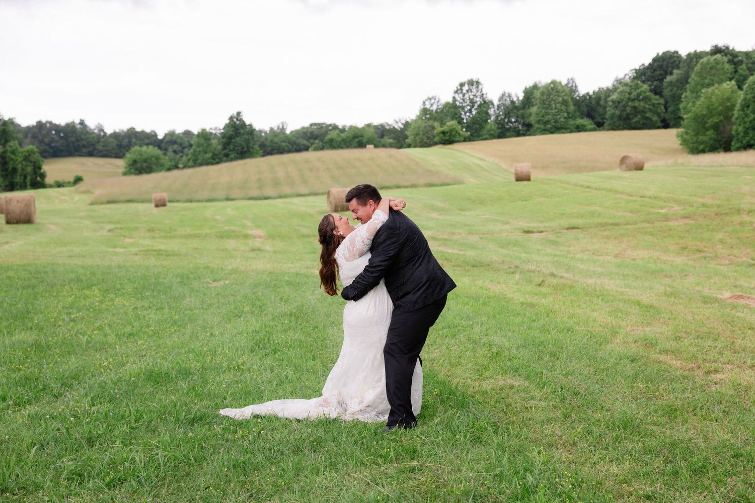 A couple dancing on a lush green hillside dotted with hay bales on a warm summer day.