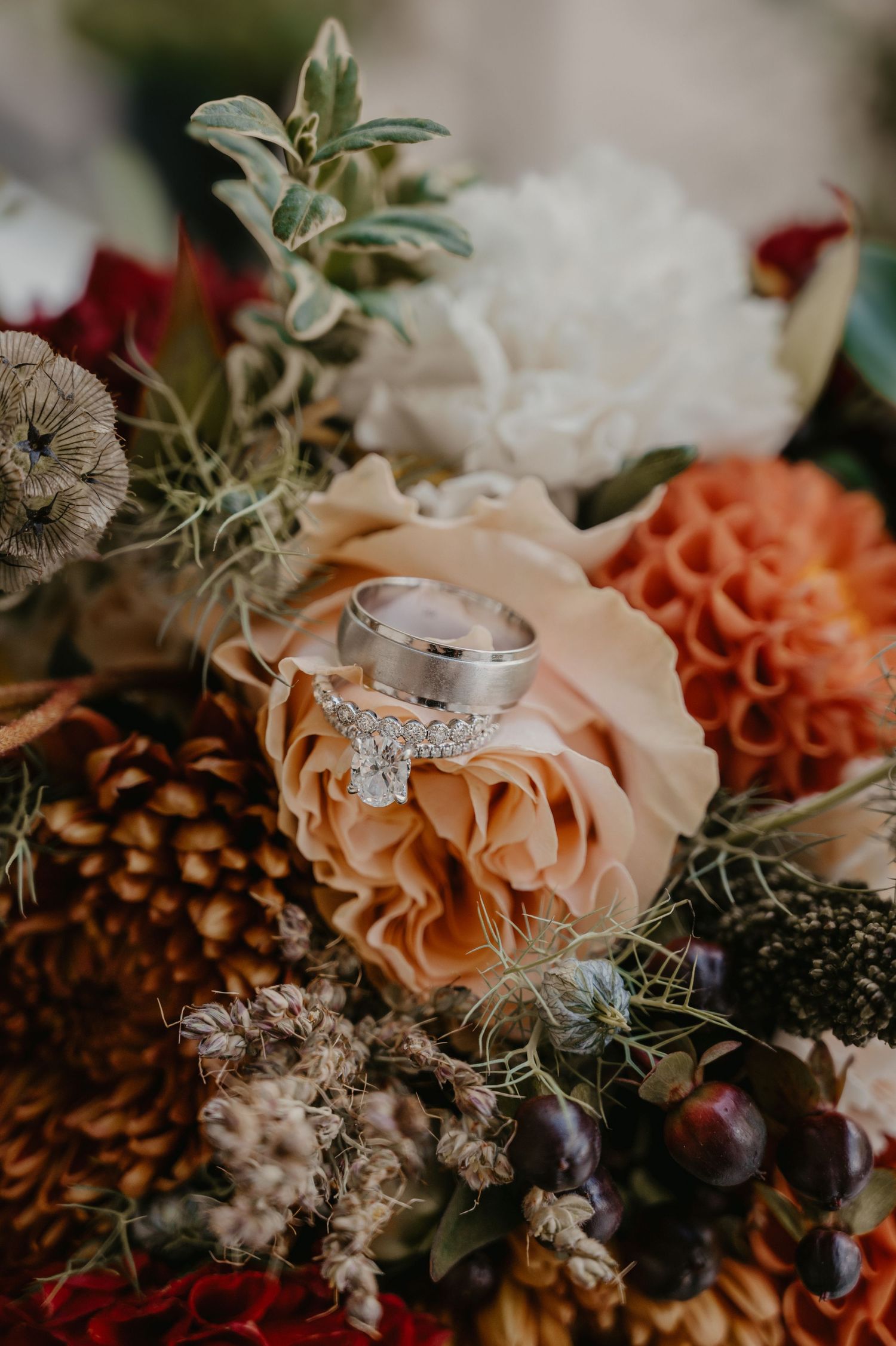 Wedding rings resting on a peach rose in a lush autumn bridal bouquet with dahlias and pinecones.