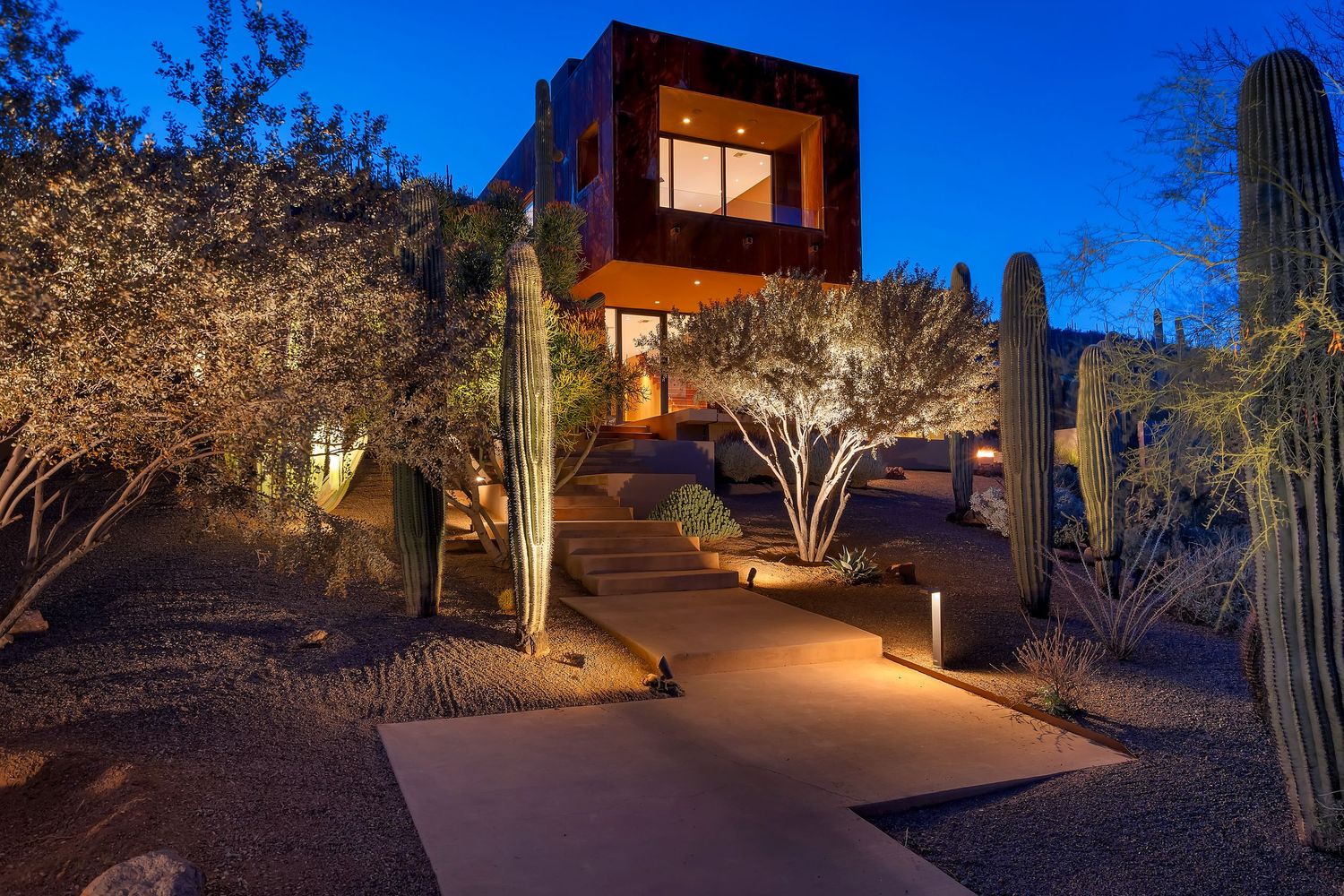 Modern desert home illuminated at dusk with saguaro cacti and pathway lighting creating a dramatic southwestern scene.