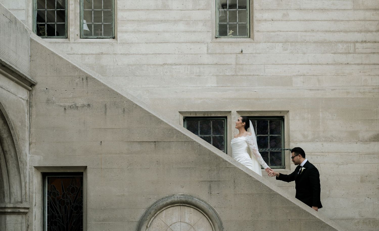 A couple in formal attire stands on an elegant stone staircase against a white building with arched windows.