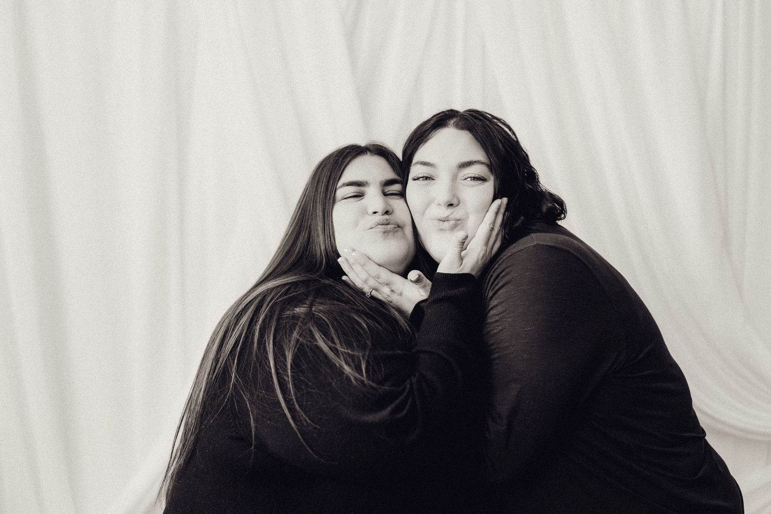 Two young women in black making kissing faces in a black and white studio portrait.