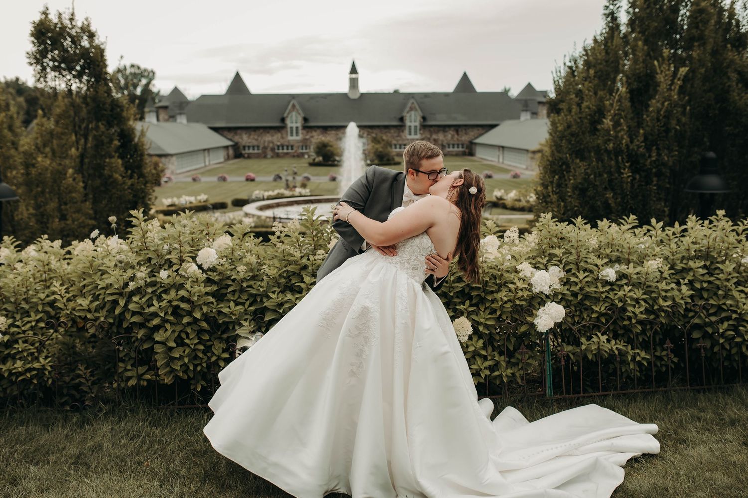 Groom dipping bride in white ball gown in lush garden at wedding venue.