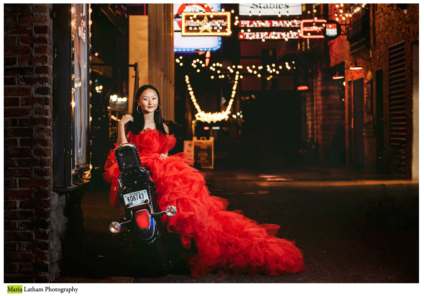 A figure in a dramatic red tulle gown poses in a dimly lit urban alleyway at night.