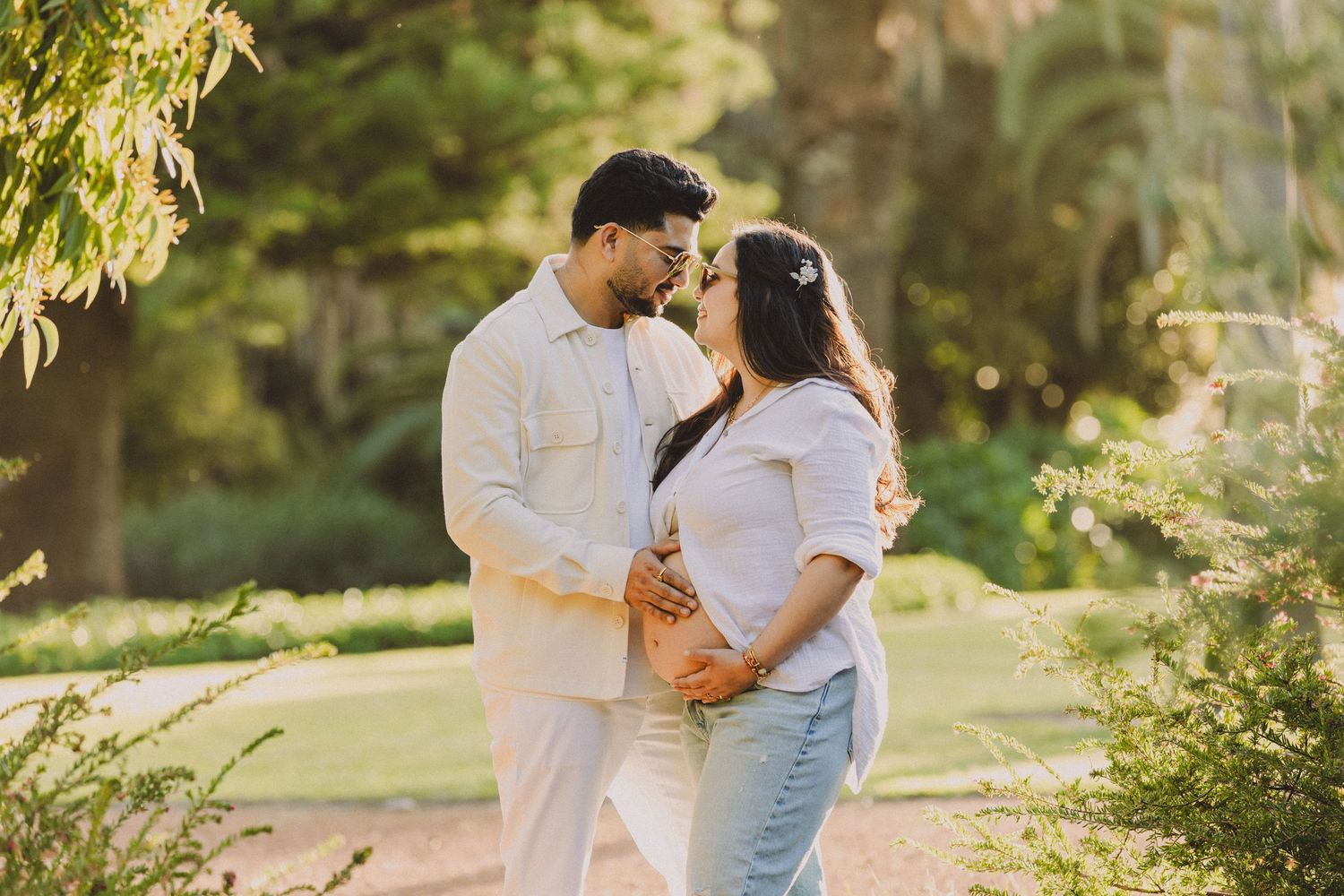 Couple in white and denim embracing outdoors in golden sunlight with lush greenery in the background.