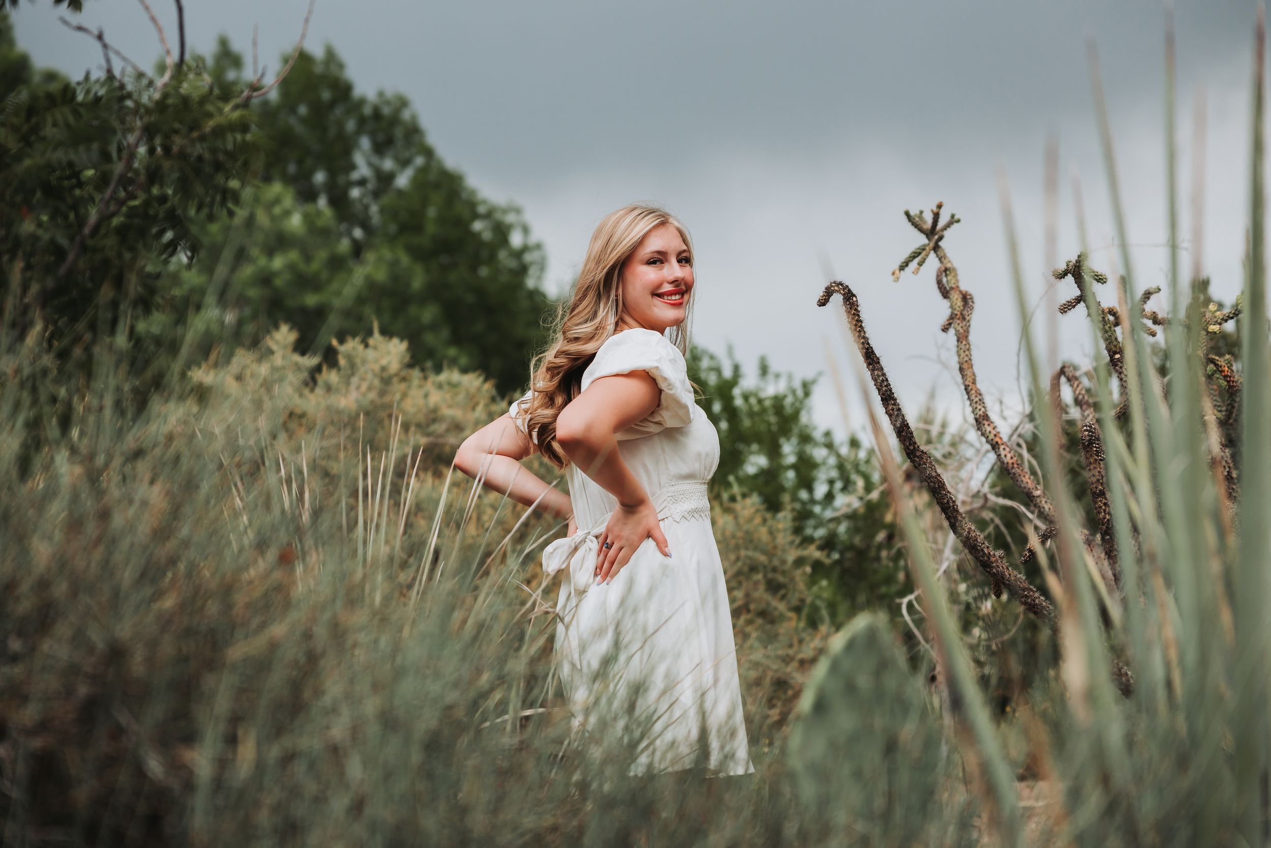 A person in a white dress poses gracefully among tall grass and dried branches in a rustic outdoor setting.