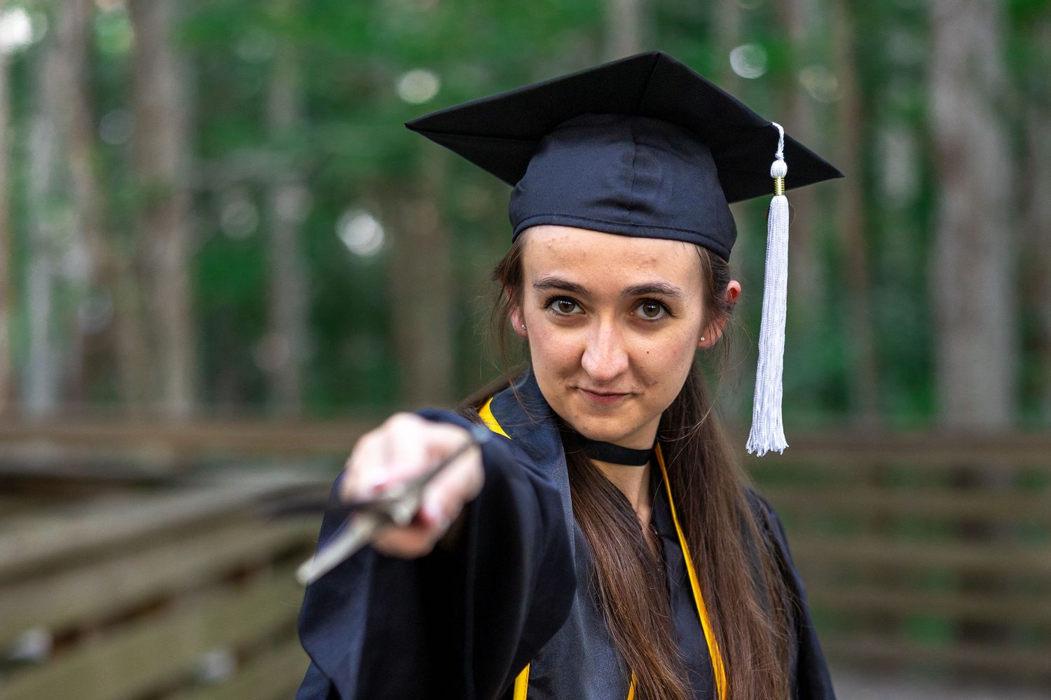 Graduate in cap and gown stands on wooden boardwalk surrounded by trees for graduation portrait photo session.