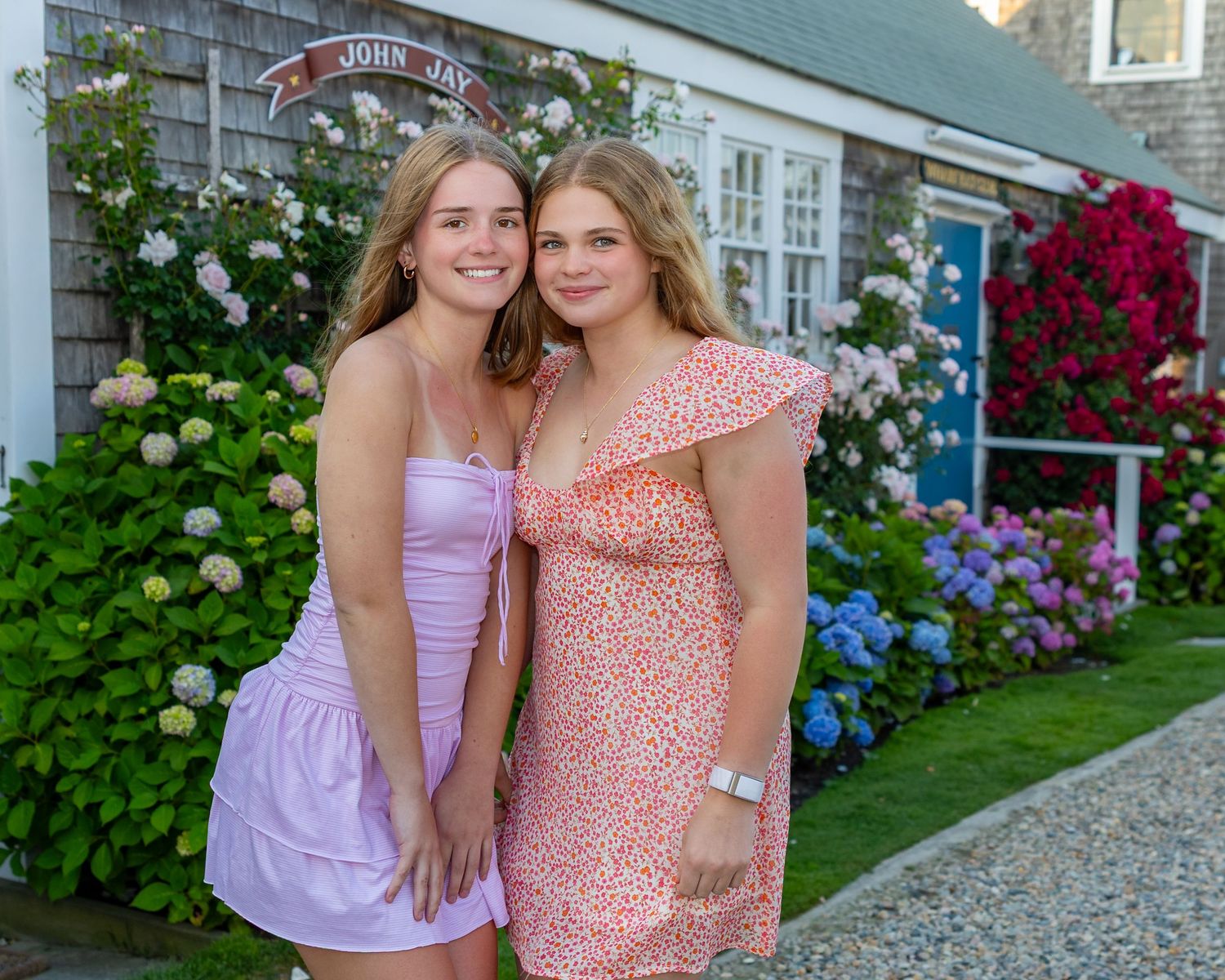 Two friends in summer dresses stand together in a garden filled with blooming hydrangeas and colorful flowers.