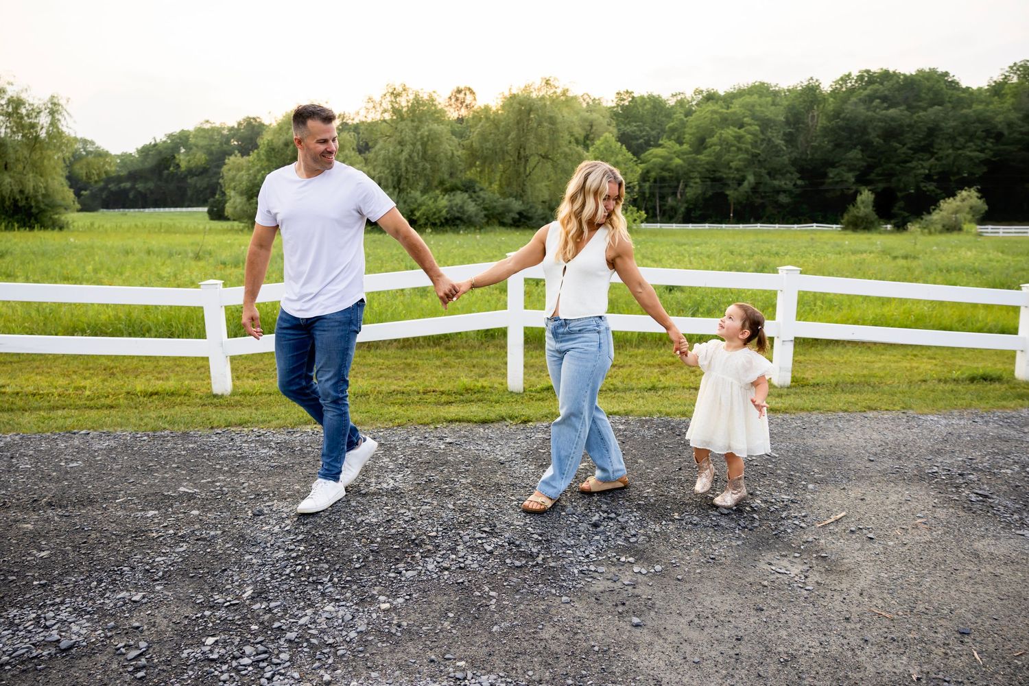 Small child in white dress watching flock of birds fly over green countryside with white fence on rural farm.