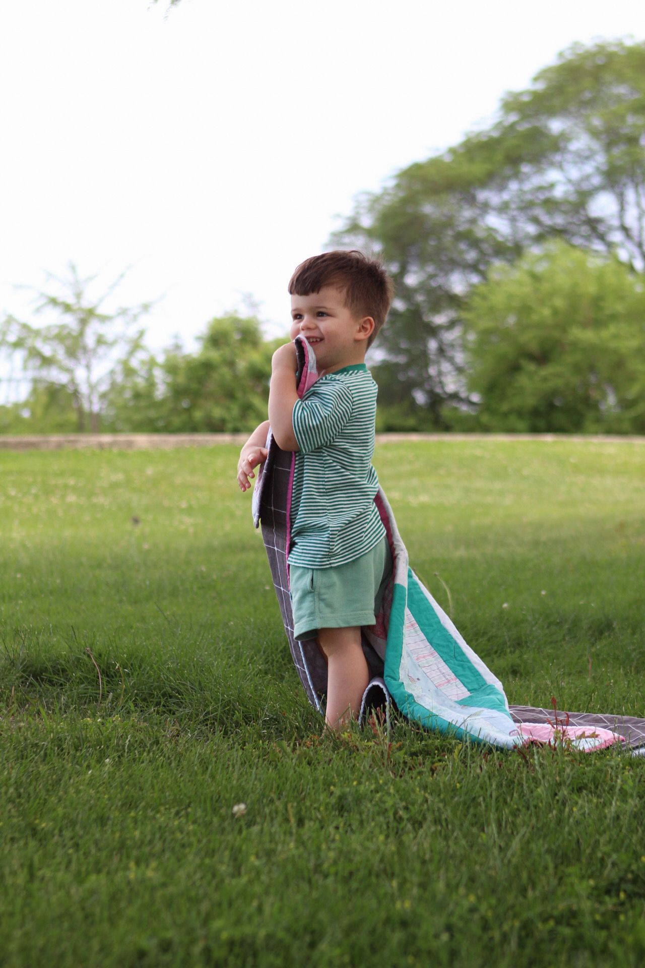 A child in a green shirt sits on a blanket in a grassy field on a sunny day.