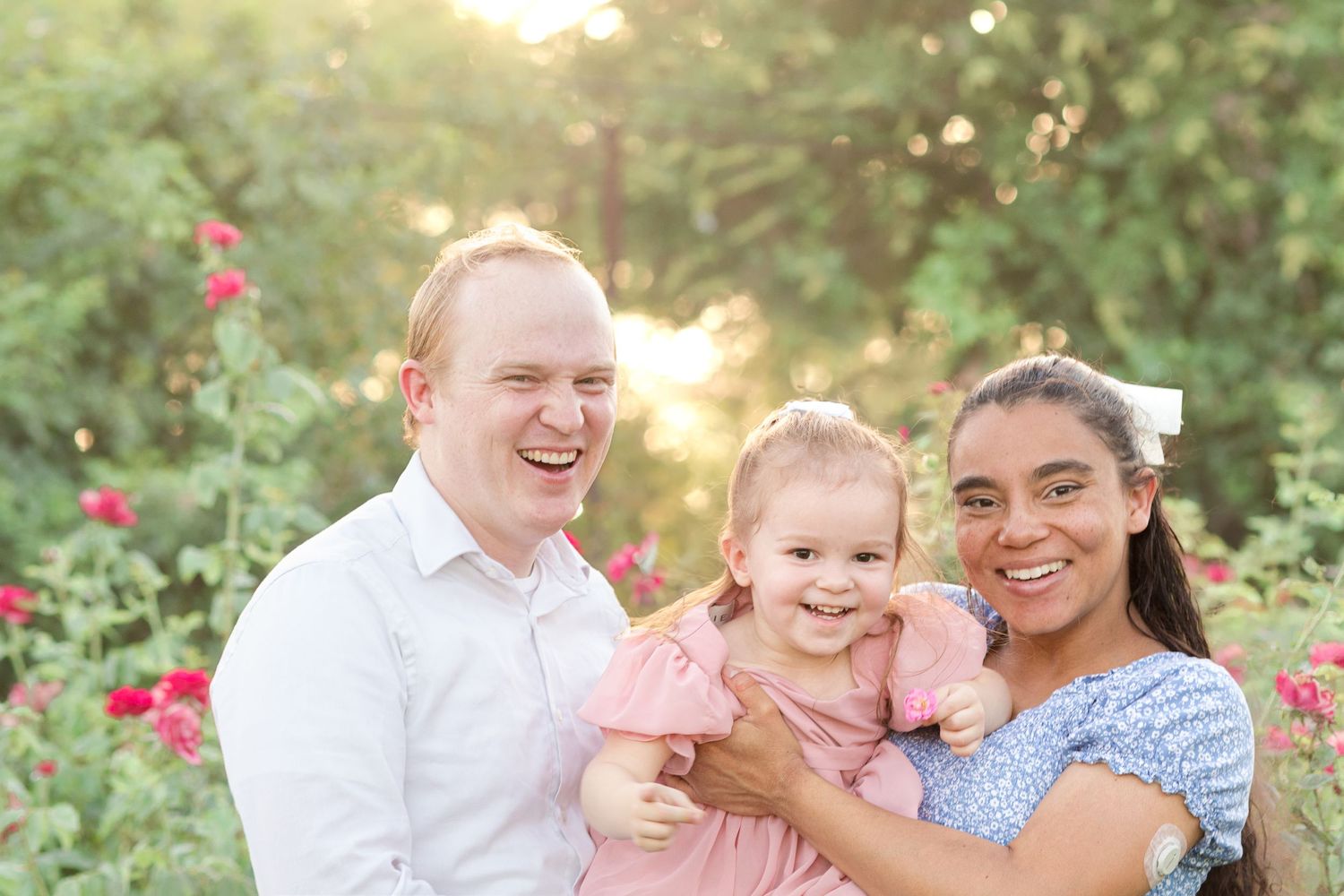 Family of 3 smiling in front of roses with the sunlight glowing through the trees behind in a Rose Garden in Mesa, Arizona.