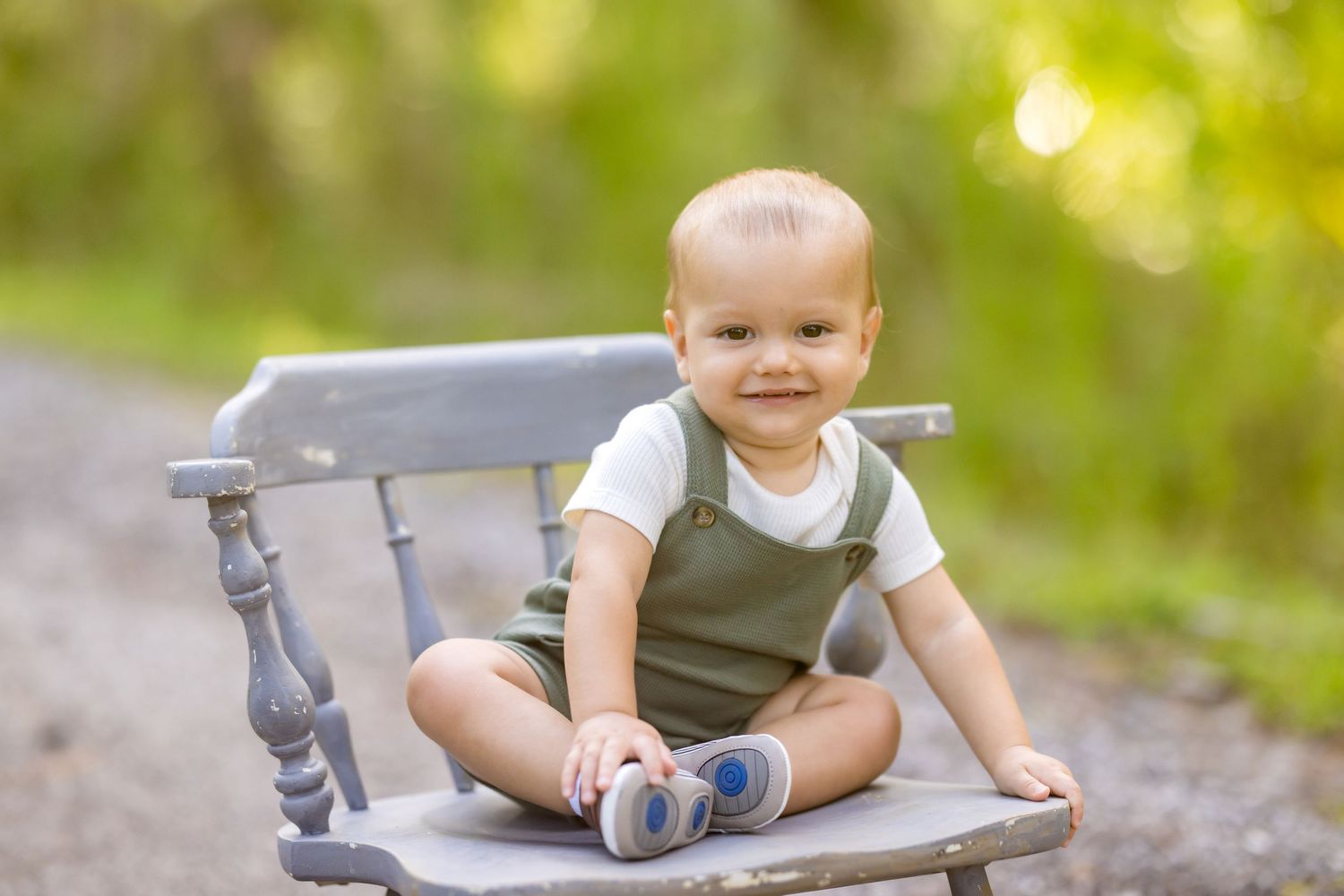 Small child in olive green overalls sits smiling on a rustic gray wooden chair outdoors with bokeh background.