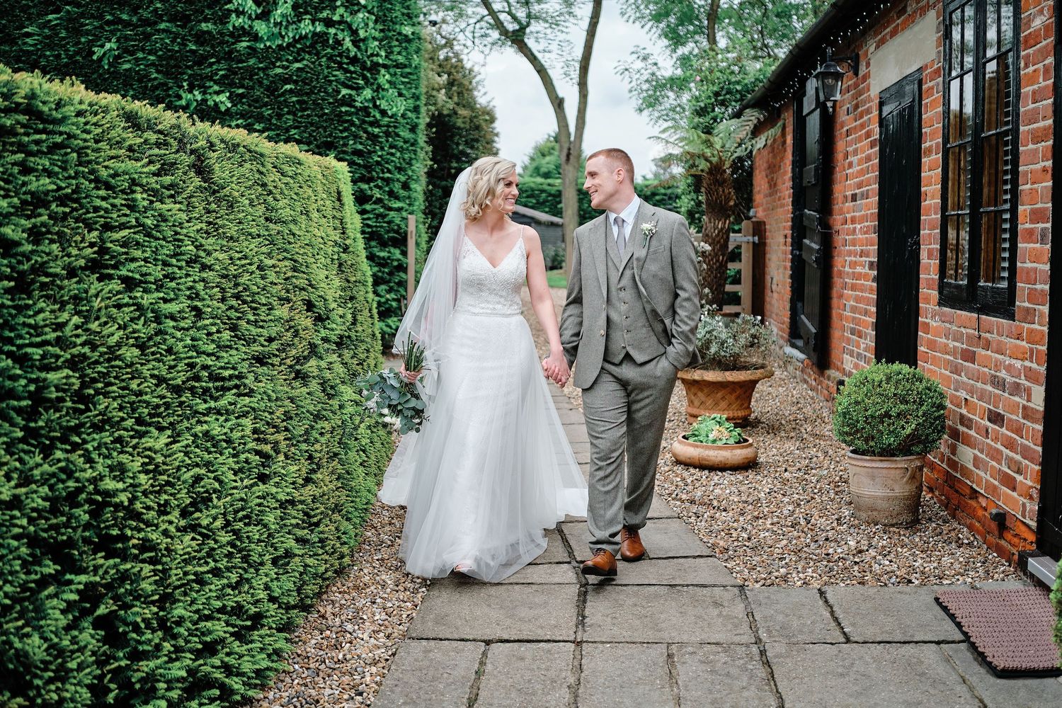 A wedding couple walks together on a brick pathway lined with lush green hedges and trees at Winters Barns wedding venue