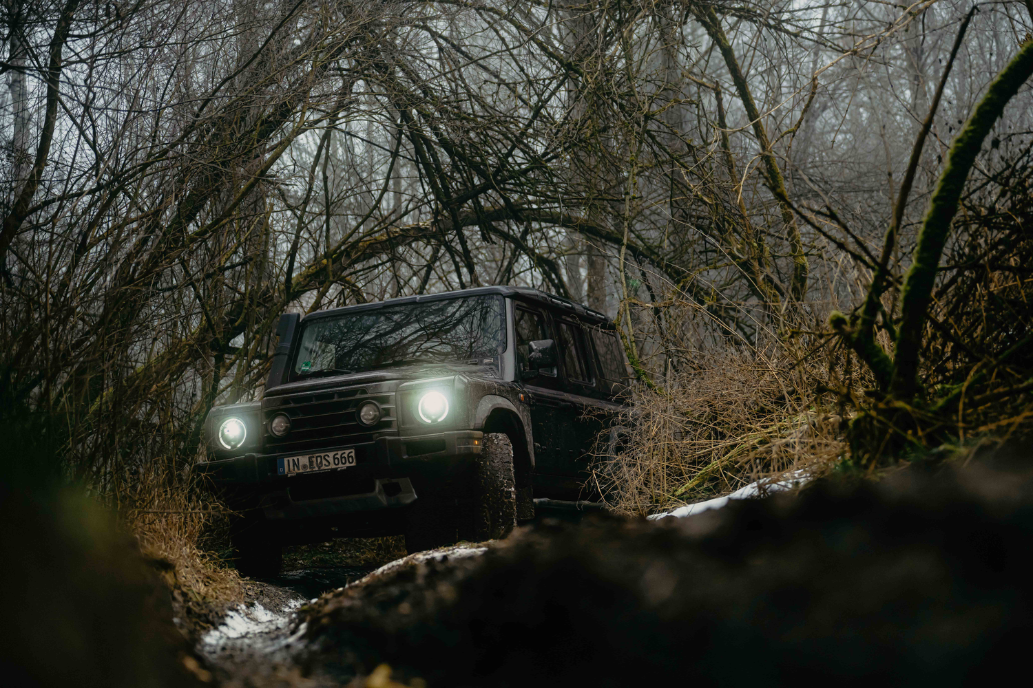 A dark Ineos Grenadier navigates through a moody forest trail with headlights illuminating the misty path.