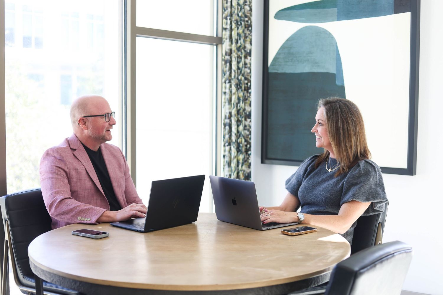 Two professionals having a business meeting at a round table with laptops in a bright modern office space.