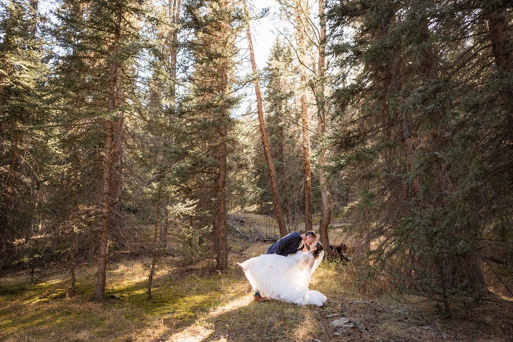A couple walks through a sunlit forest path during their outdoor wedding photo session in the mountains.