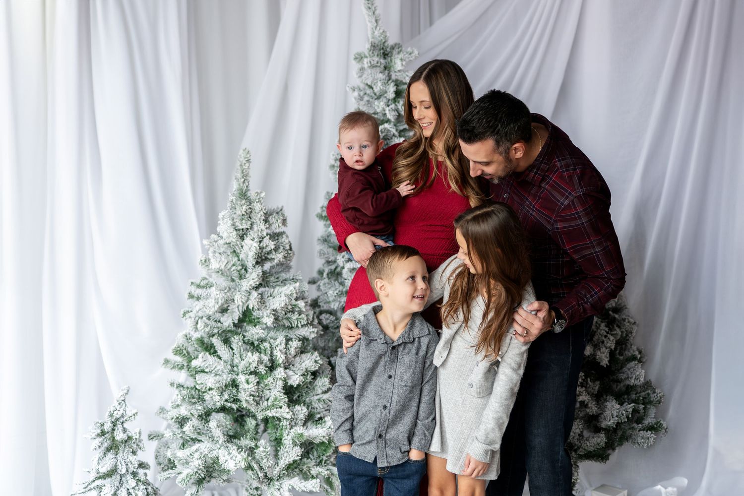 Family of five in red and plaid holiday outfits poses near flocked white Christmas trees in a studio.