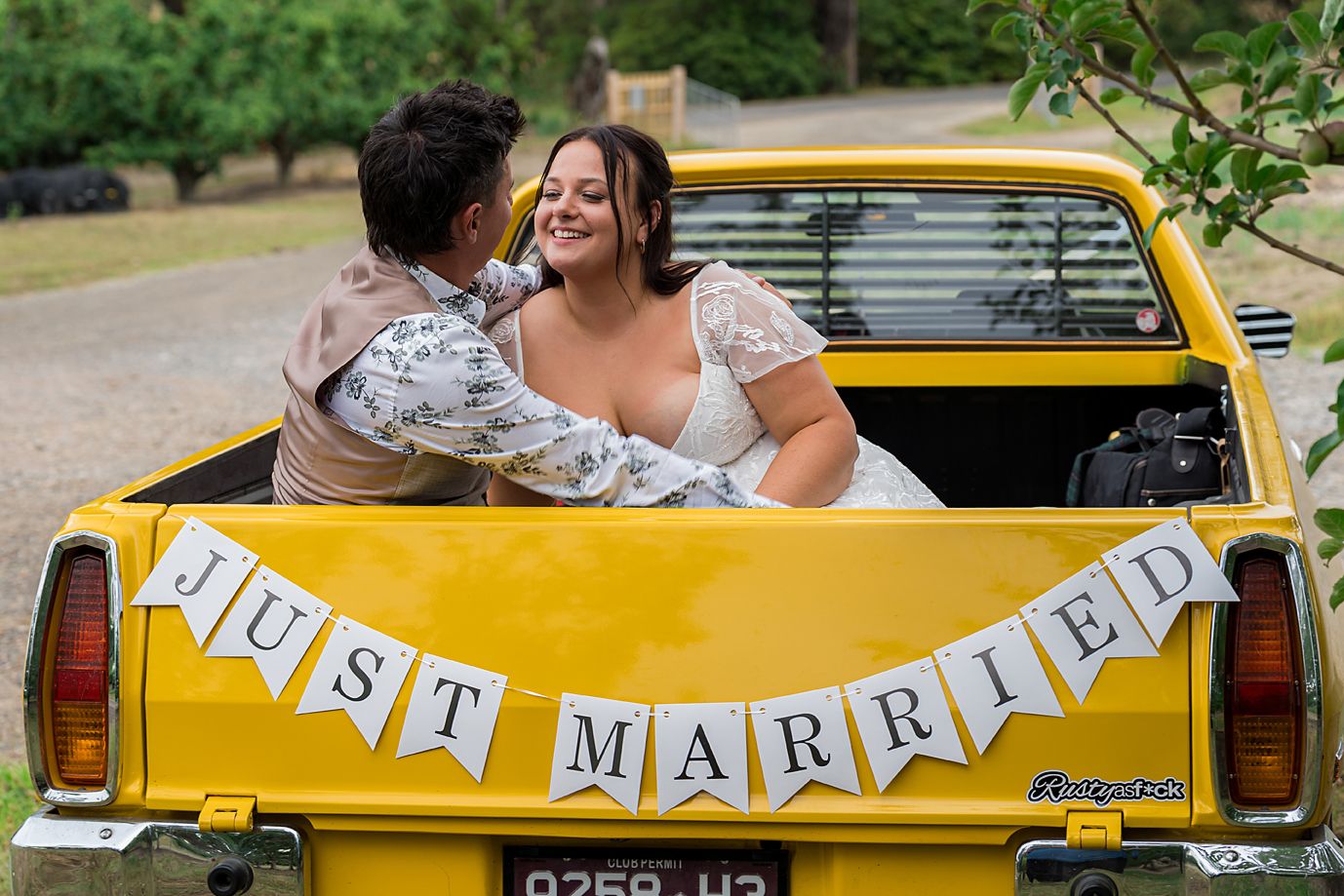 A yellow vintage car decorated with 'Just Married' bunting banner drives through a lush green park setting.