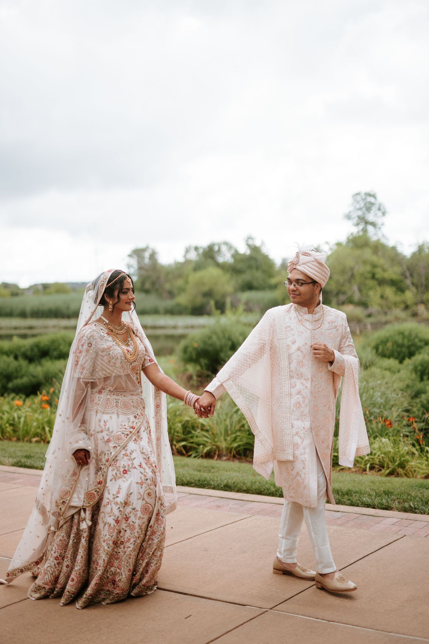 A couple in traditional South Asian wedding attire walks and laughs together on a paved outdoor path.