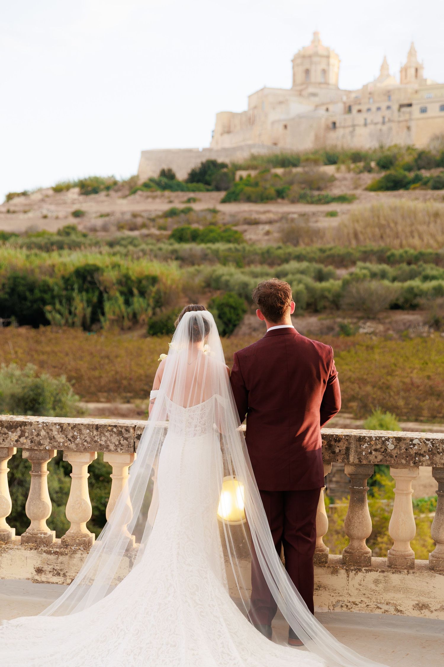 Newly married couple walking along brick path by white picket fence under tree canopy.