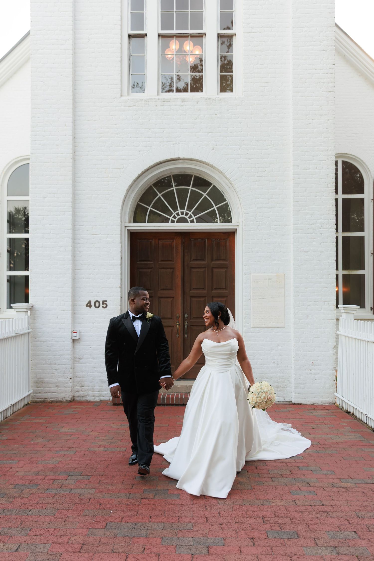 Newly married couple walking along brick path by white picket fence under tree canopy.