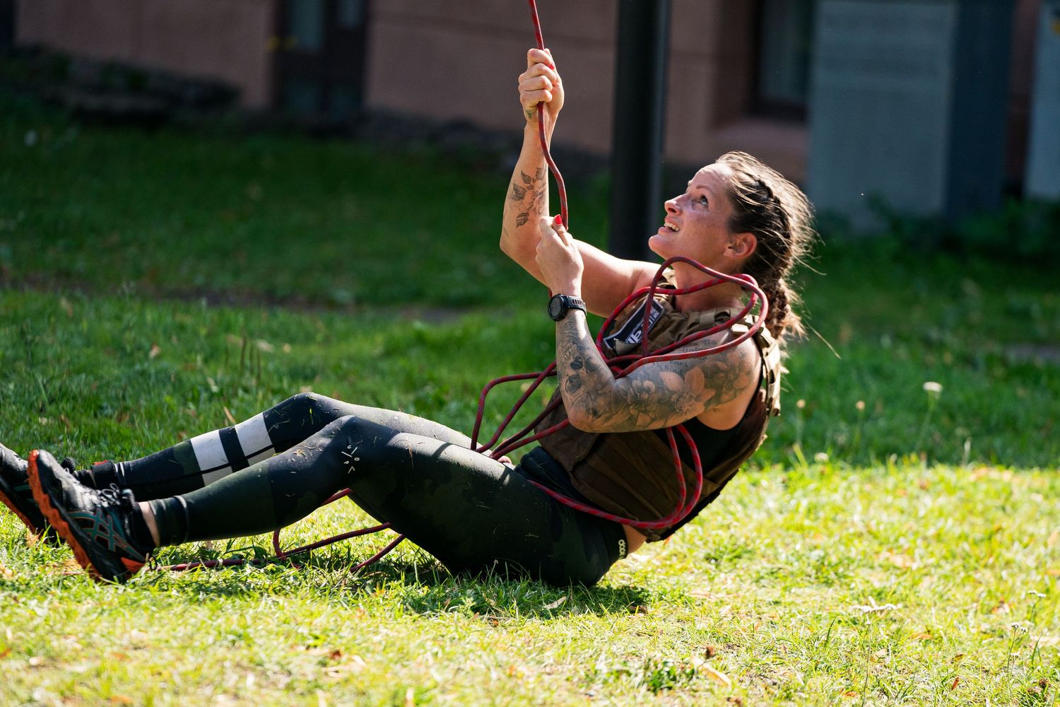 A person wearing a leather vest slides down a zip line on a sunny day in a grassy area.
