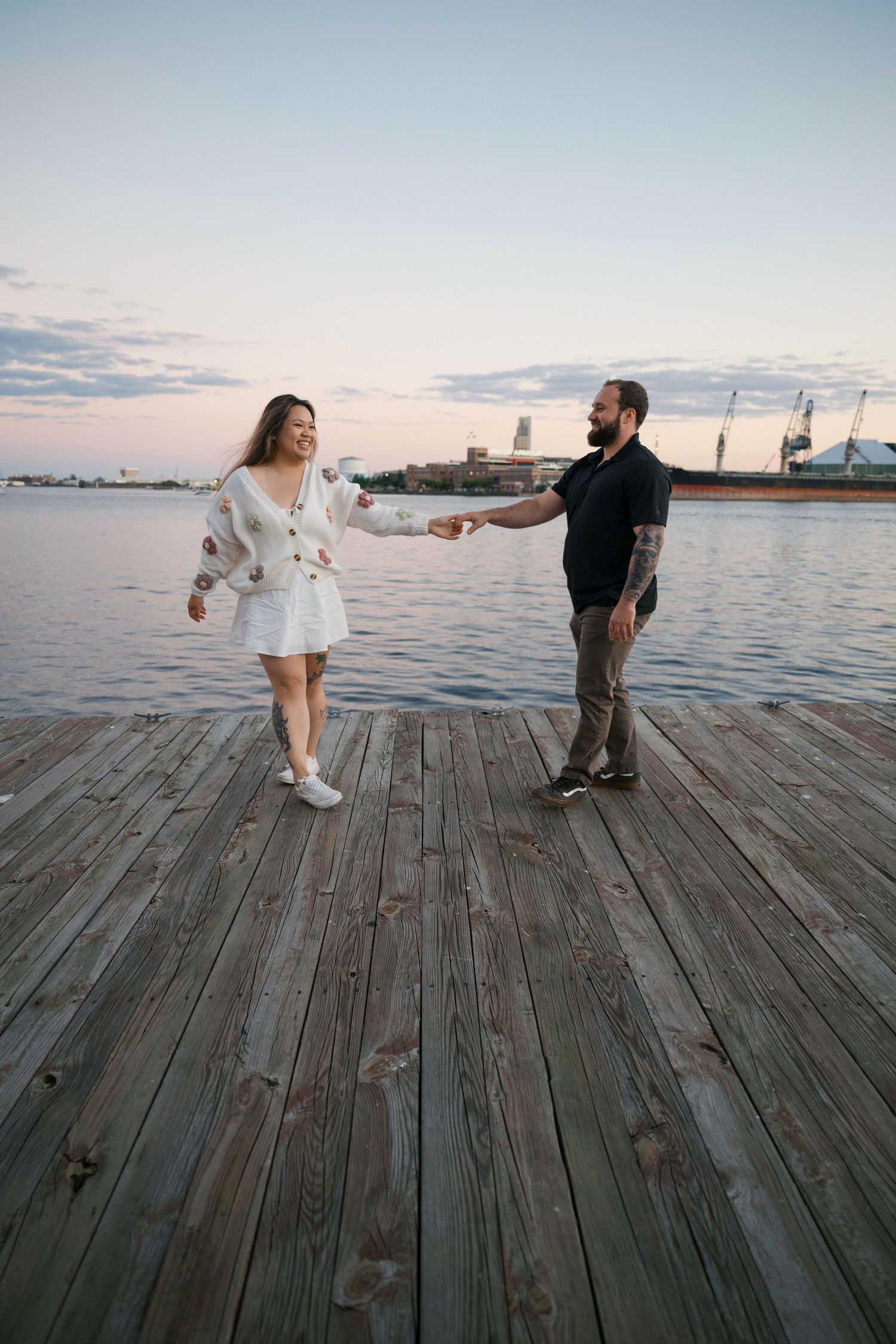 Couple dancing in sequence on a wooden pier by the waterfront at sunset with a cityscape in the background.