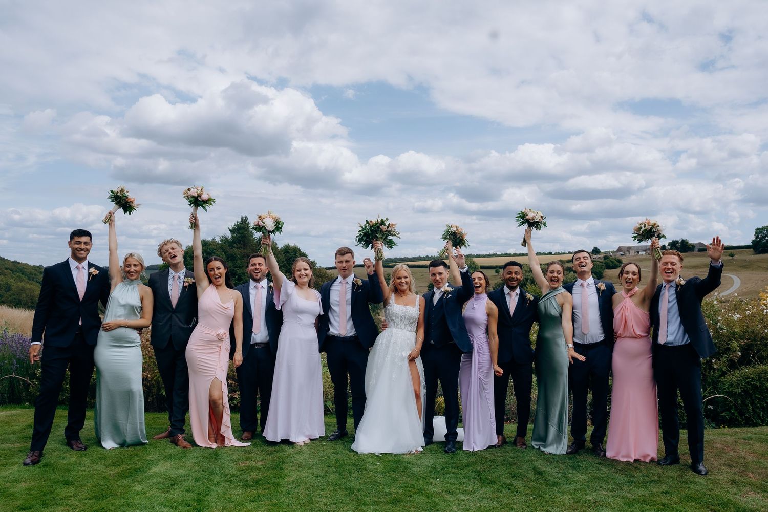 Wedding party in formal attire poses together outdoors with pink bridesmaid dresses against a cloudy sky backdrop.