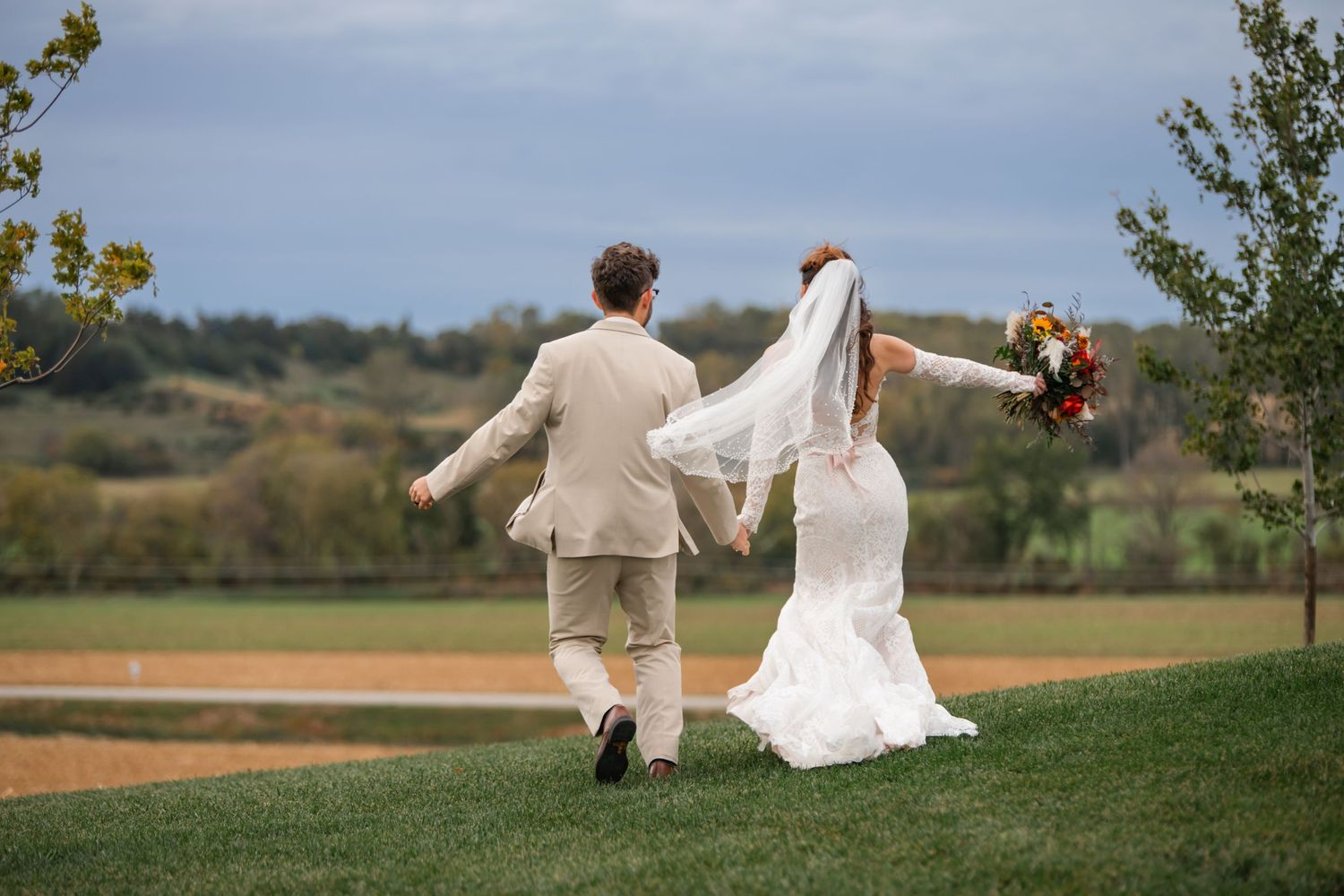 A couple in wedding attire walks hand in hand across a grassy hilltop overlooking scenic countryside.