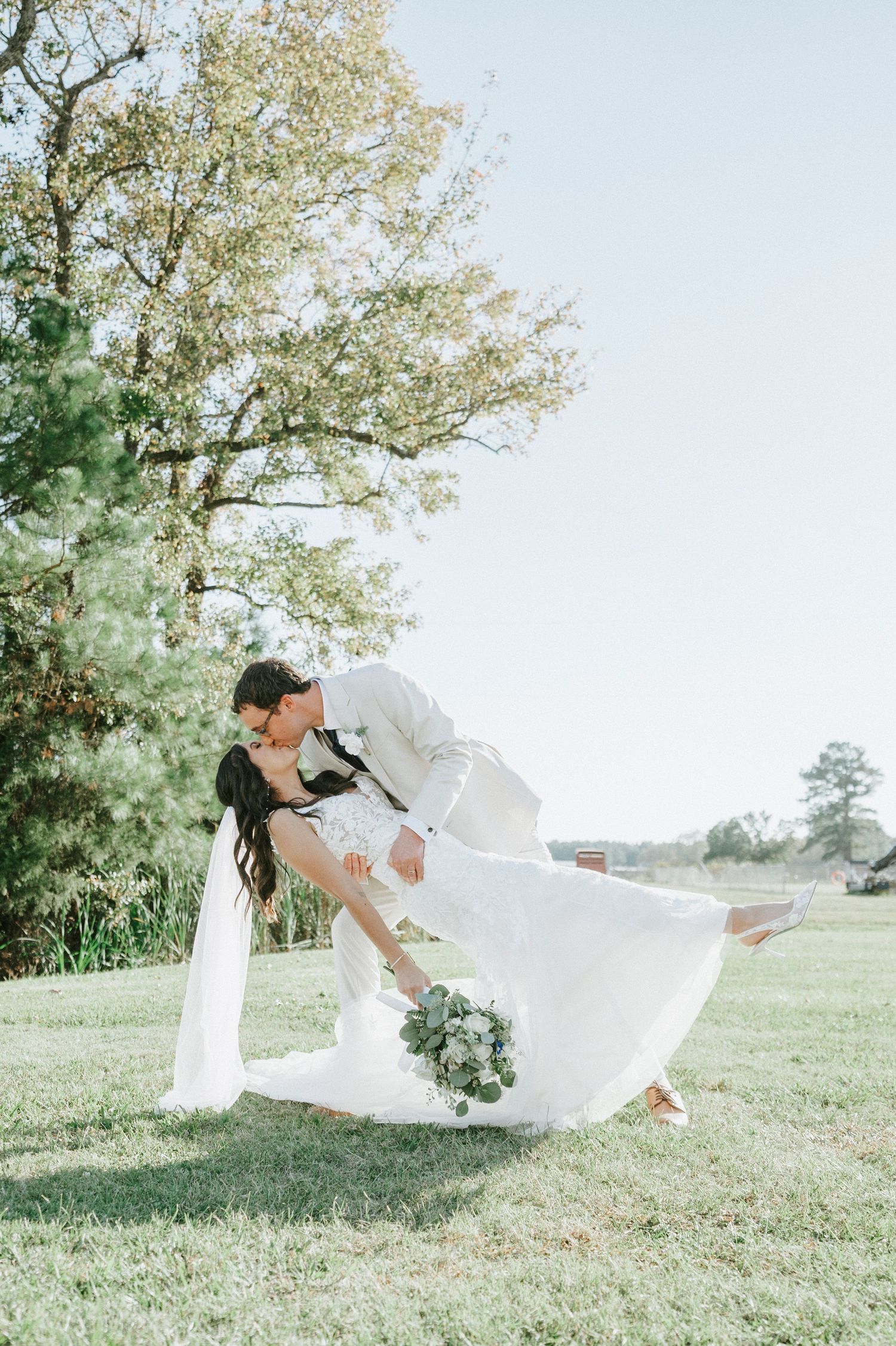 A couple shares a romantic dip kiss pose in a scenic outdoor setting with green grass and trees in the background.