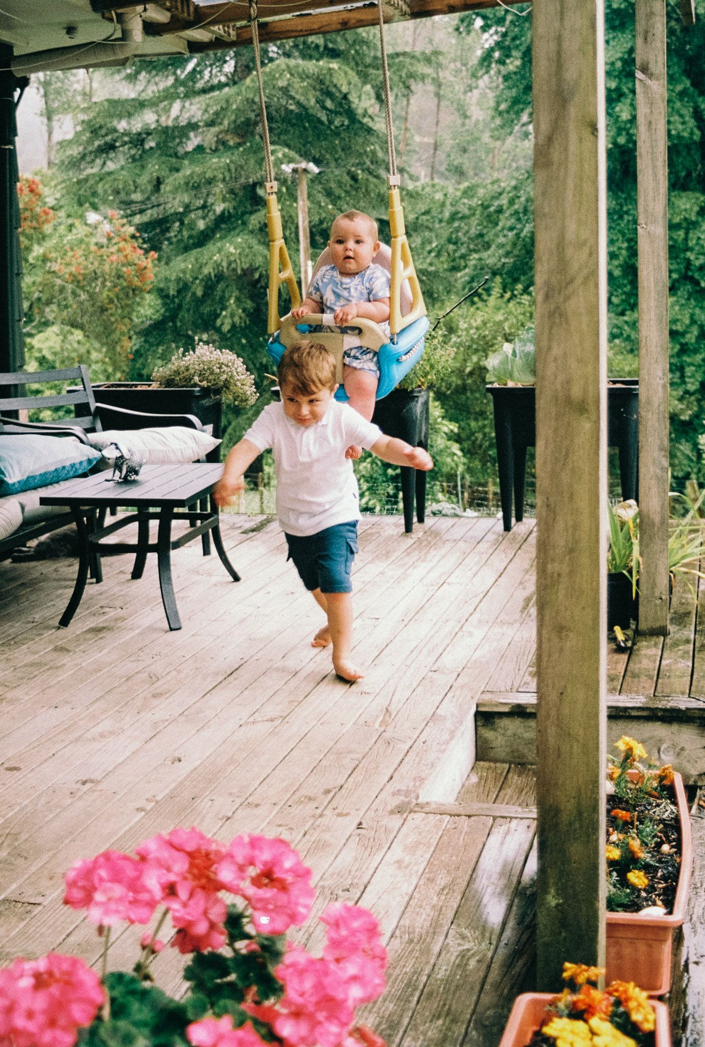 A child swings joyfully on a wooden deck surrounded by blooming flowers and tall trees on a sunny day.