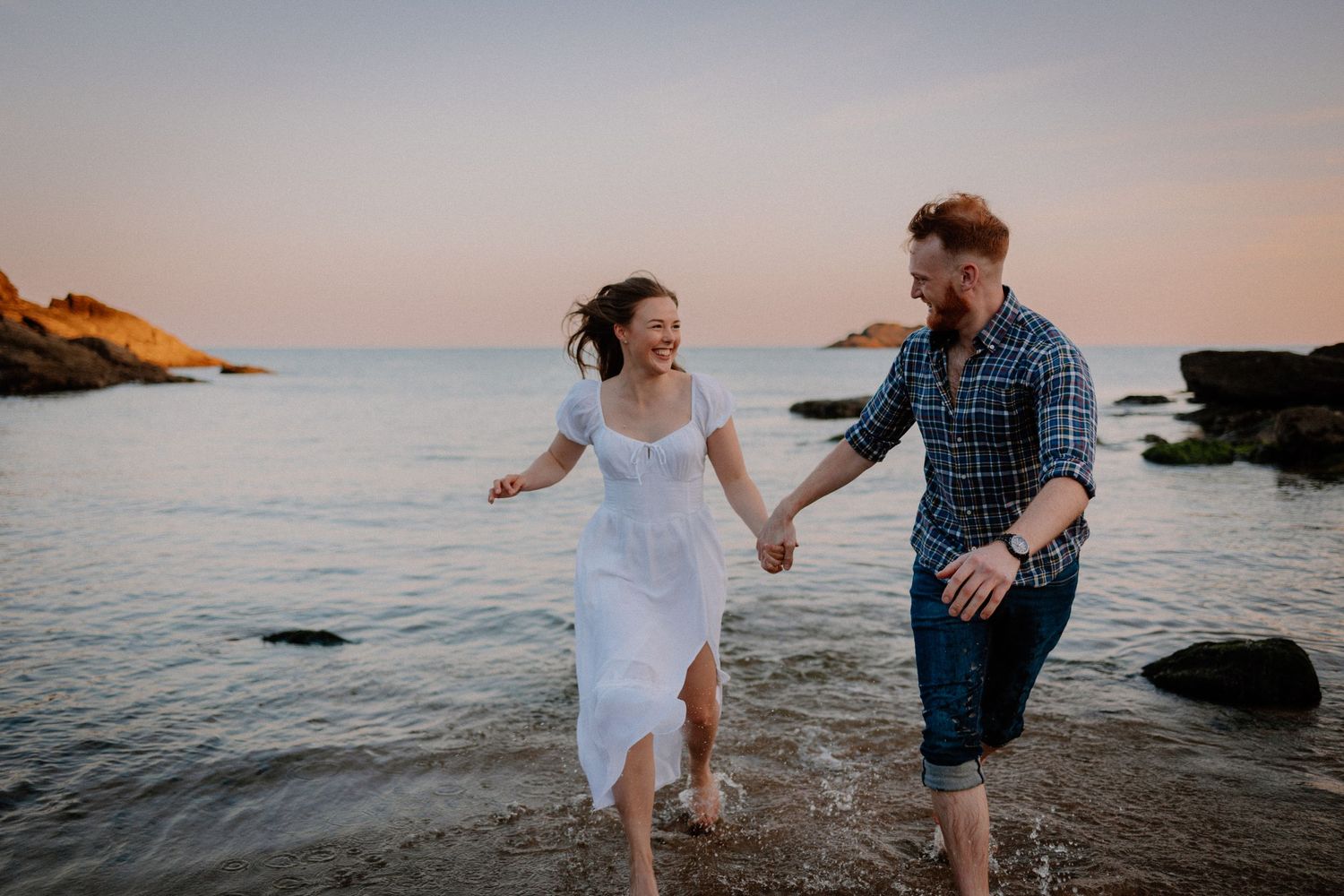 A couple in white and blue attire playfully runs and splashes along the beach shoreline at sunset.