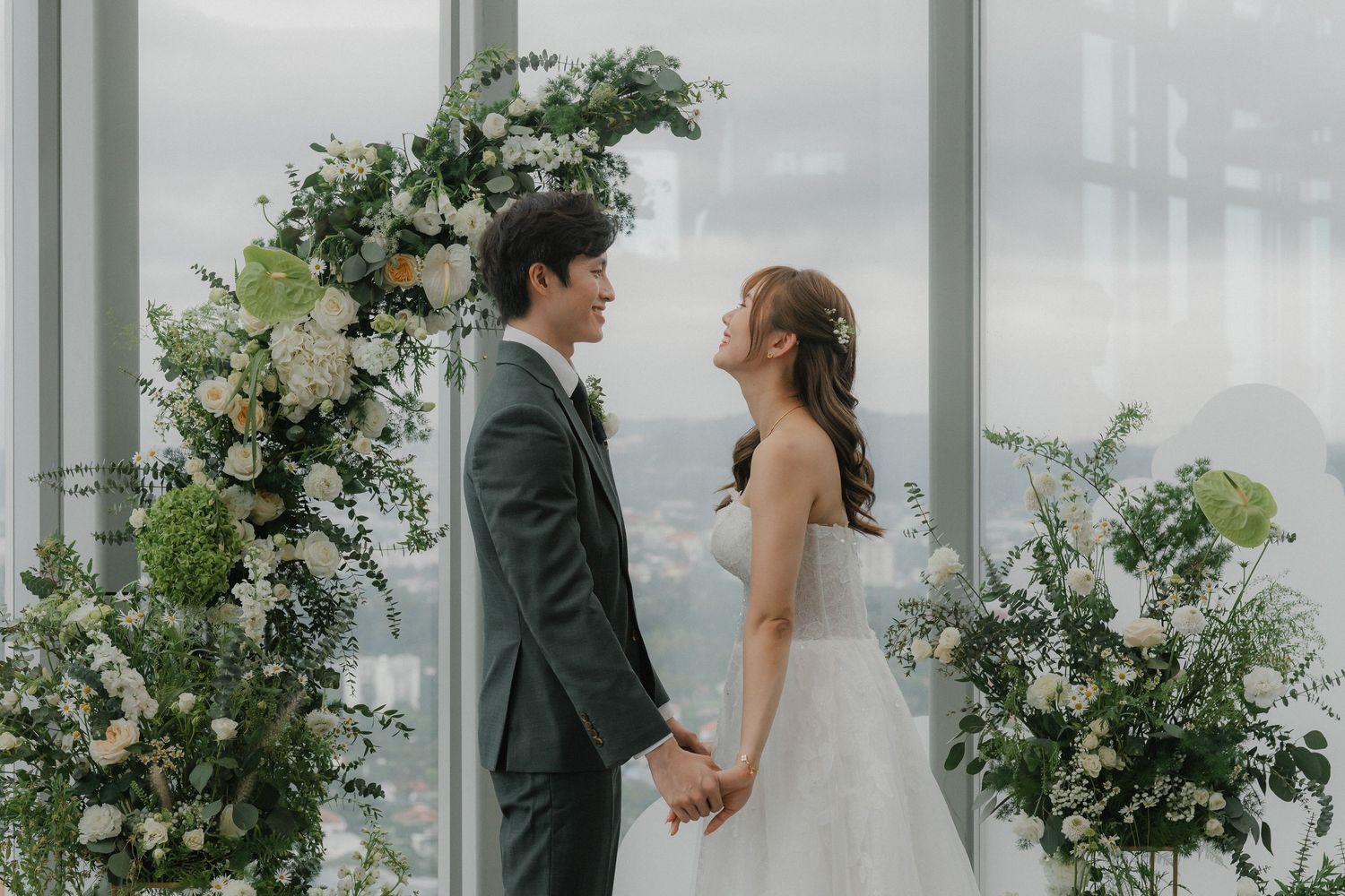 A romantic wedding ceremony setup with greenery and floral arrangements in front of large windows overlooking a cloudy sky.