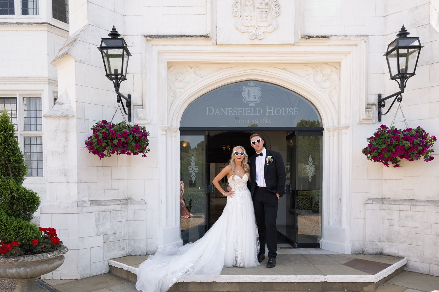 Elegant wedding couple enters through ornate doorway with hanging flower baskets and lanterns at stone building entrance.