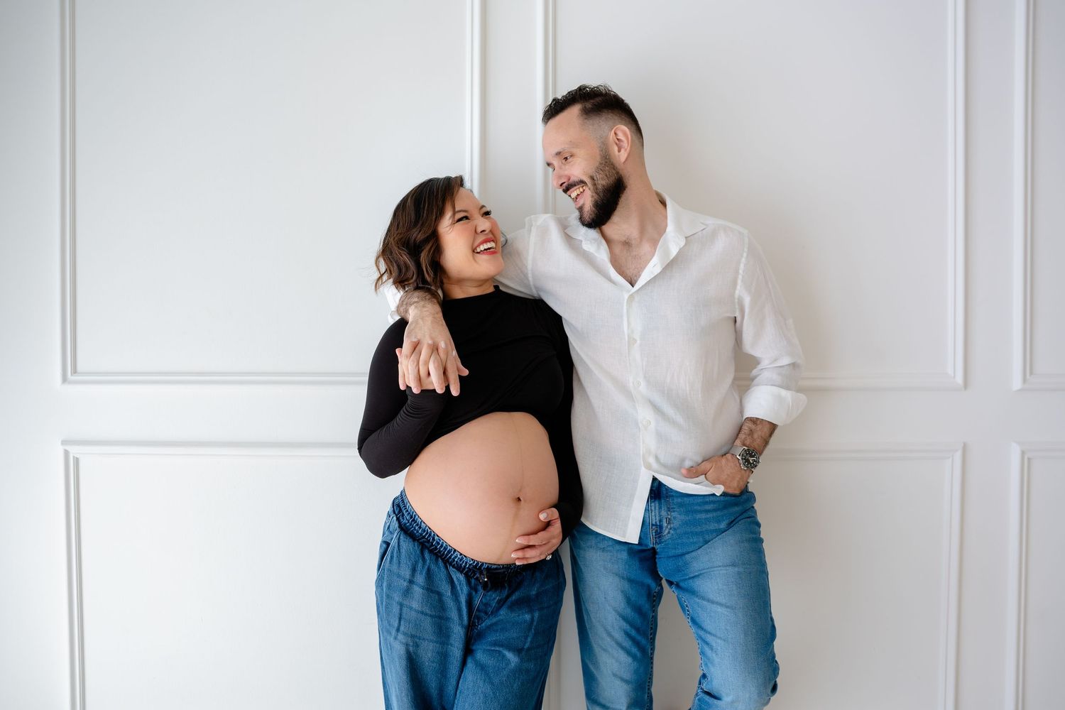 A couple shares intimate moments during a romantic indoor photoshoot against a white wall.