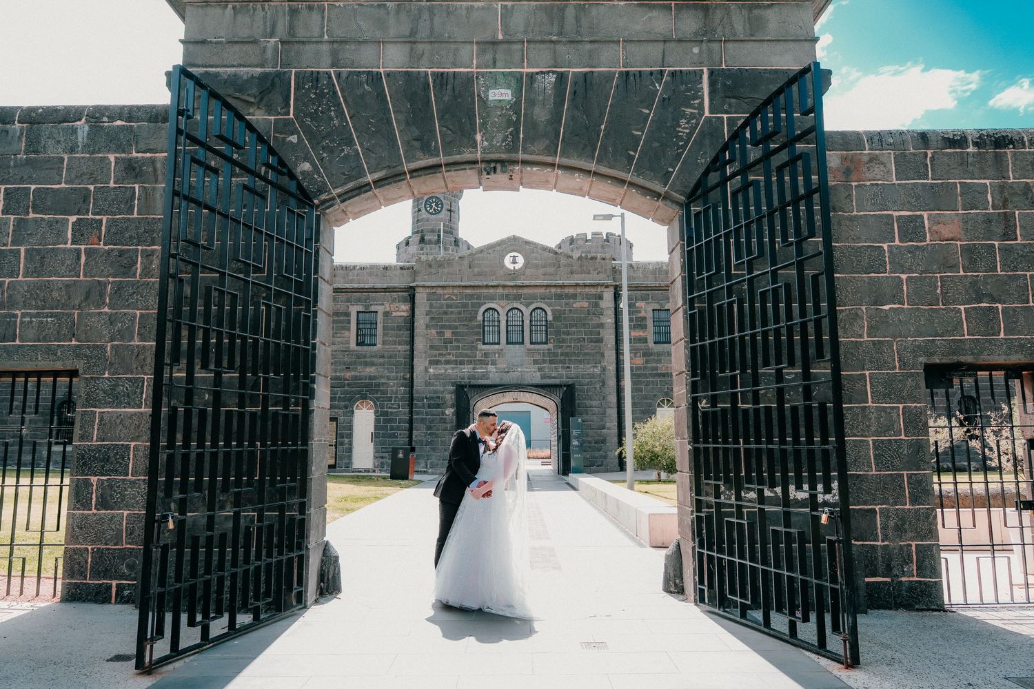 A wrought iron gate entrance to a historic stone building with a clock tower against a bright blue sky.