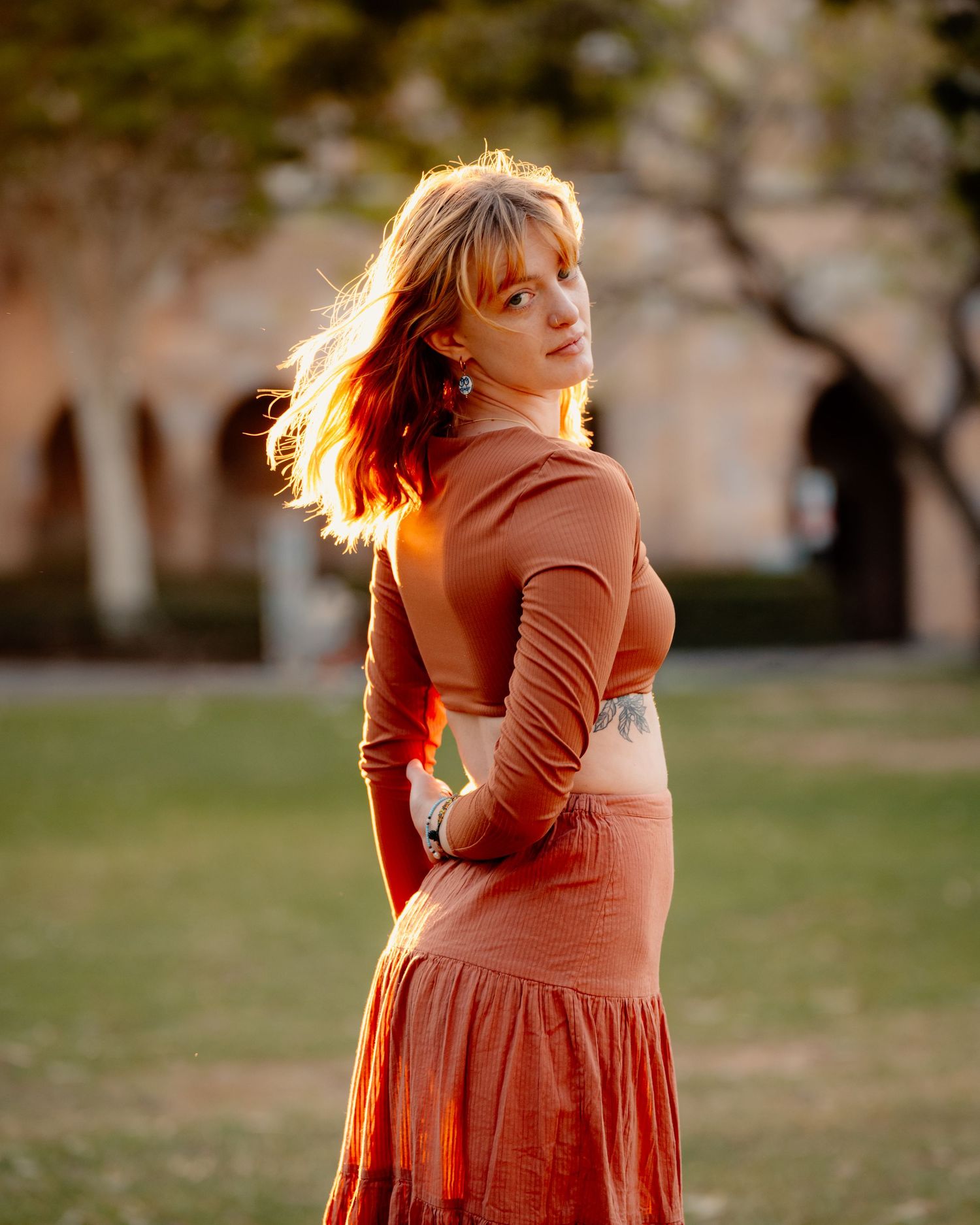 A woman in a rust-colored dress poses playfully in golden sunlight during an outdoor maternity photoshoot.