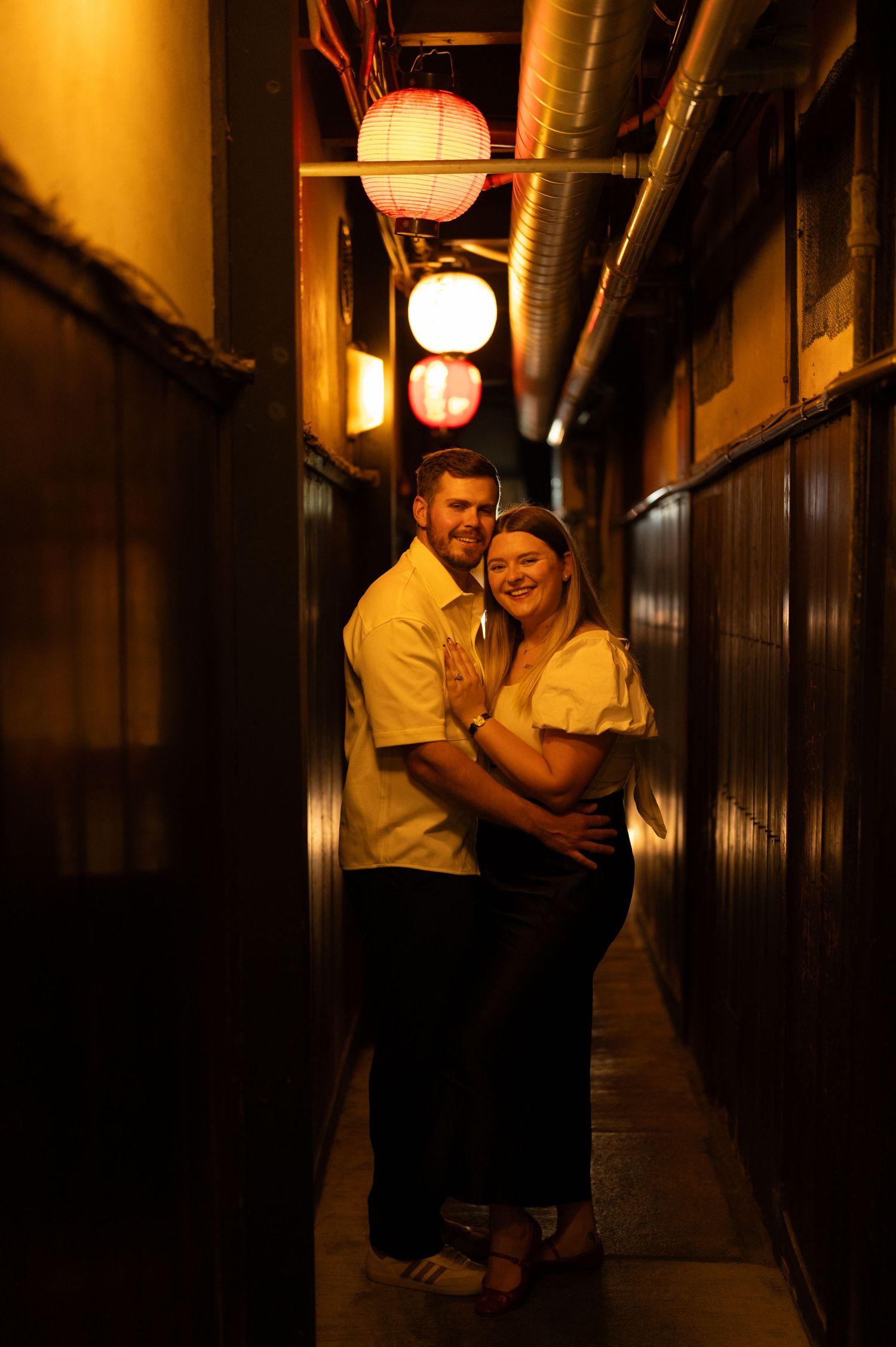 Happy couple hugging in a narrow lantern-lit Japanese alleyway at night.