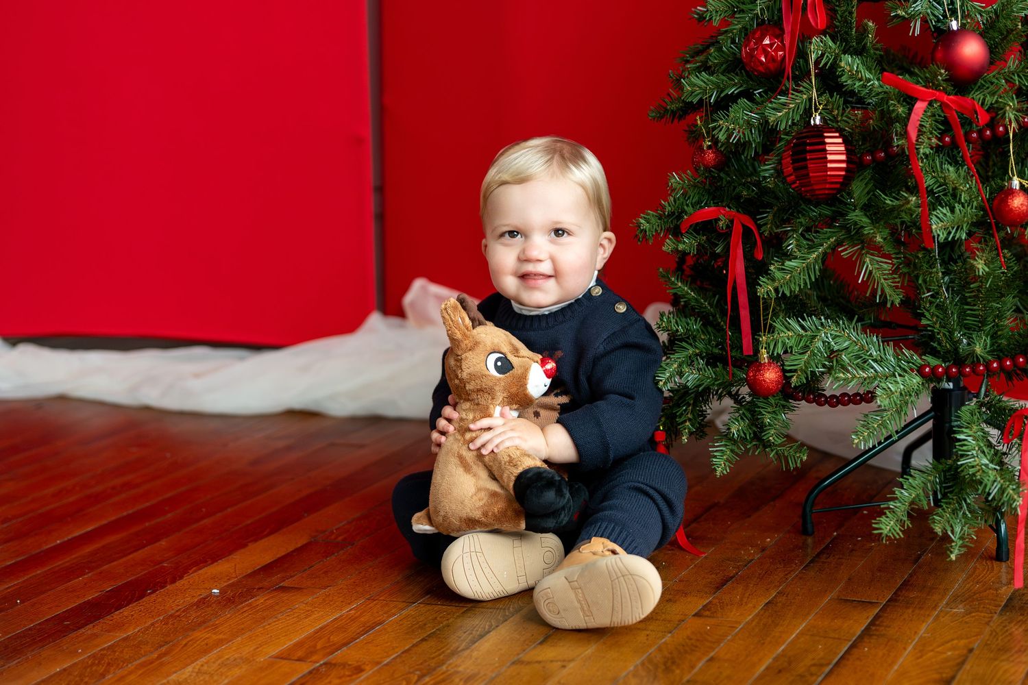 Smiling toddler in navy reindeer sweater holds stuffed Rudolph toy by Christmas tree.