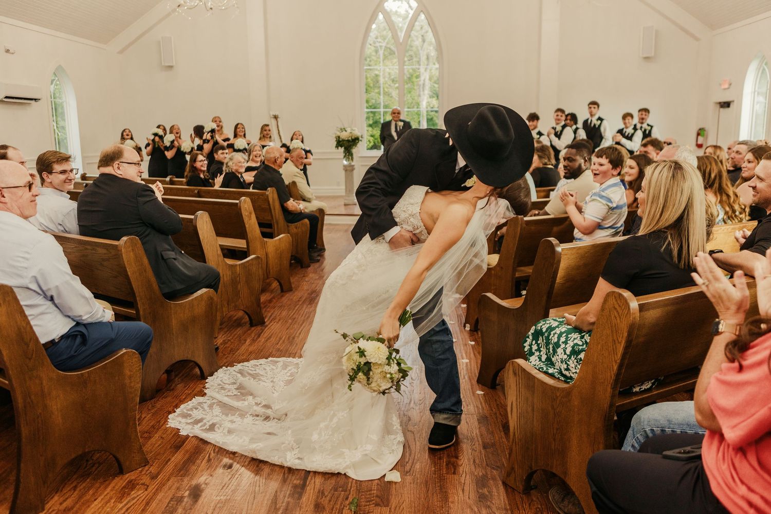 A romantic kiss between newlyweds in formal wedding attire against a bright outdoor backdrop.