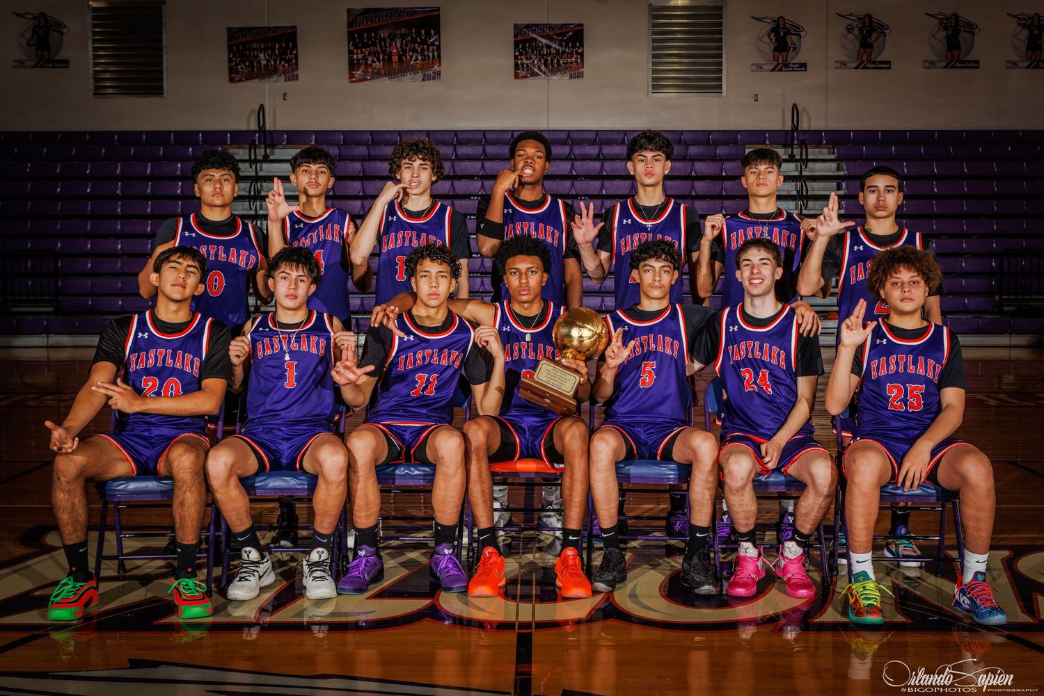 Basketball players in purple jerseys sit on a bench during team photo session in a gymnasium.