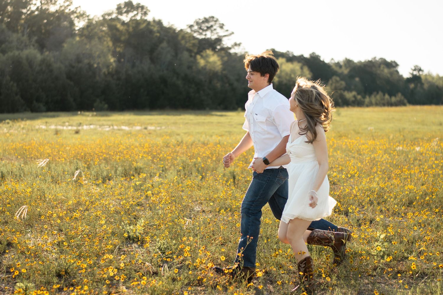 A couple runs playfully through a sunlit field of yellow wildflowers.
