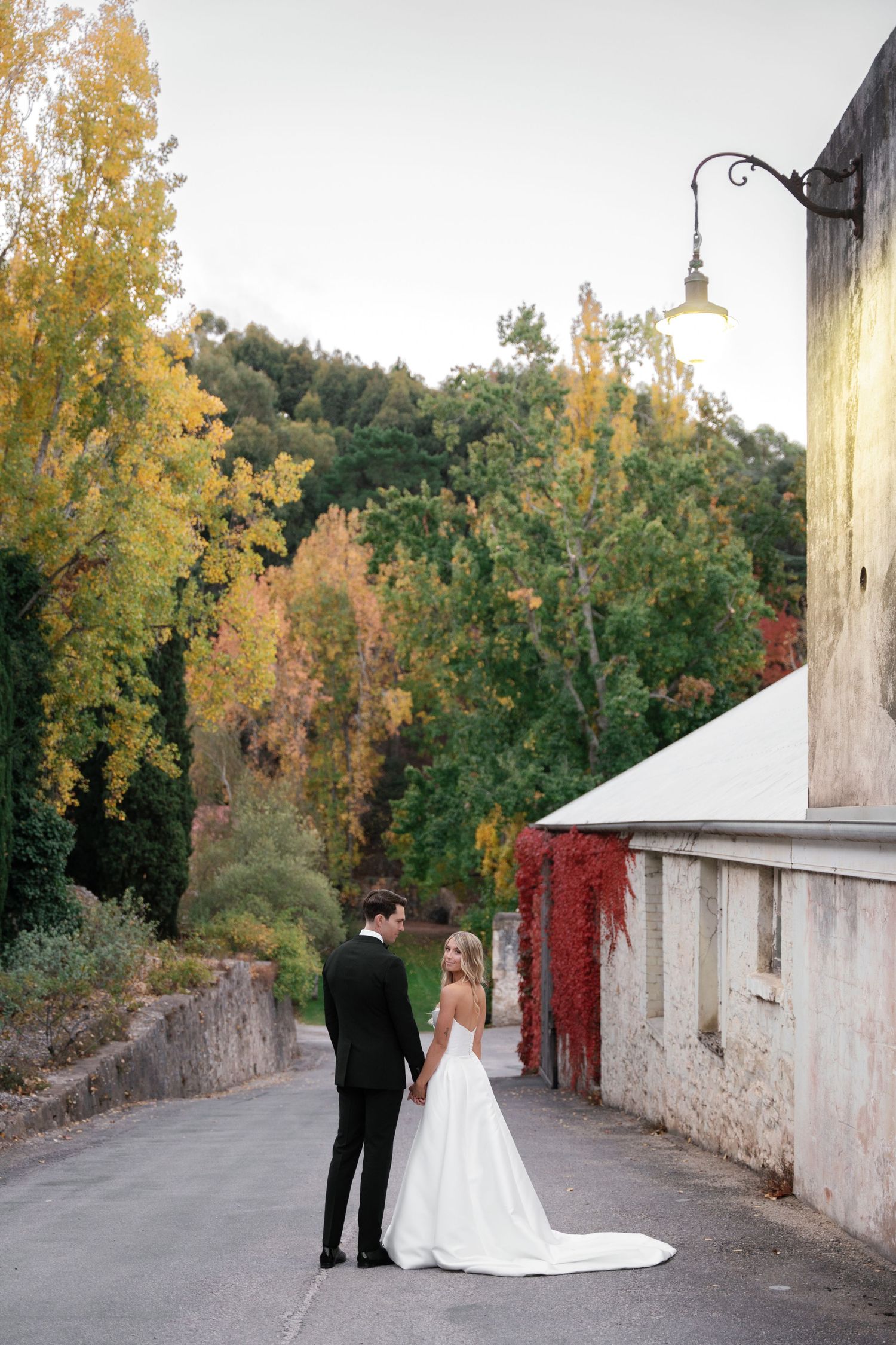 Newlyweds share quiet moment on autumn path with historic stone wall and fall foliage.
