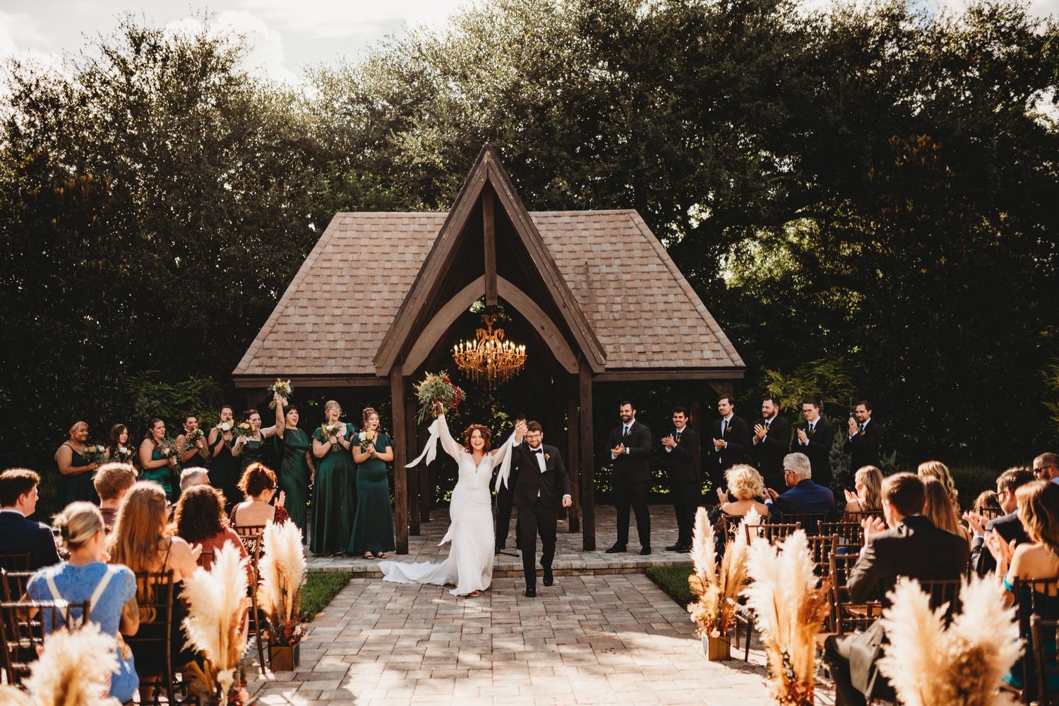 A wedding ceremony takes place at a rustic wooden chapel with guests seated on both sides of an outdoor aisle.