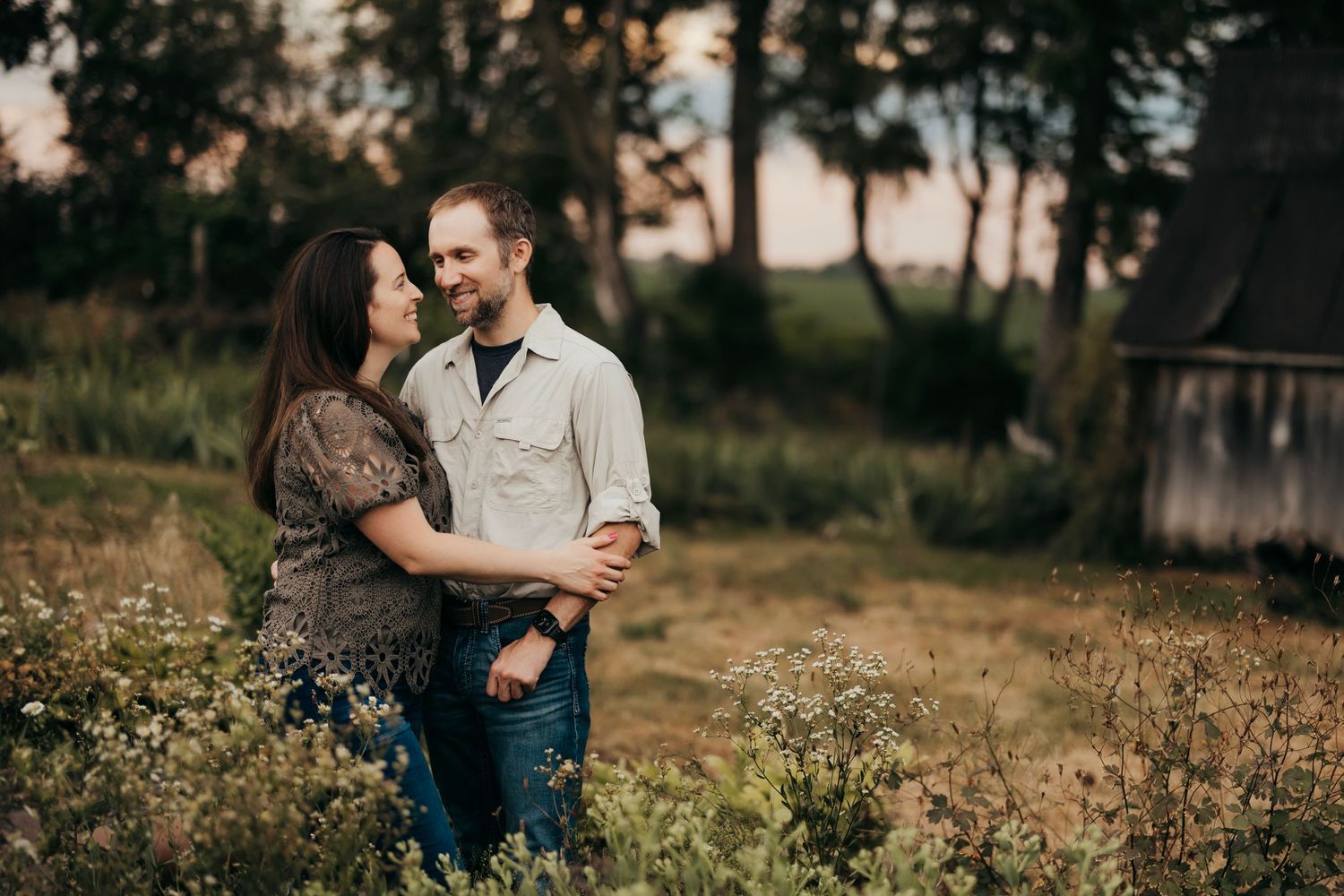 Couple walks hand in hand through a golden sunset field near rustic barn.