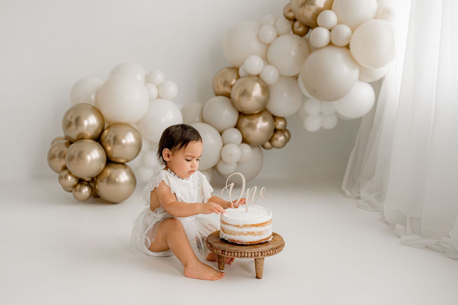 Baby in white outfit sits by birthday cake with gold and white balloon arch decoration in studio setting.